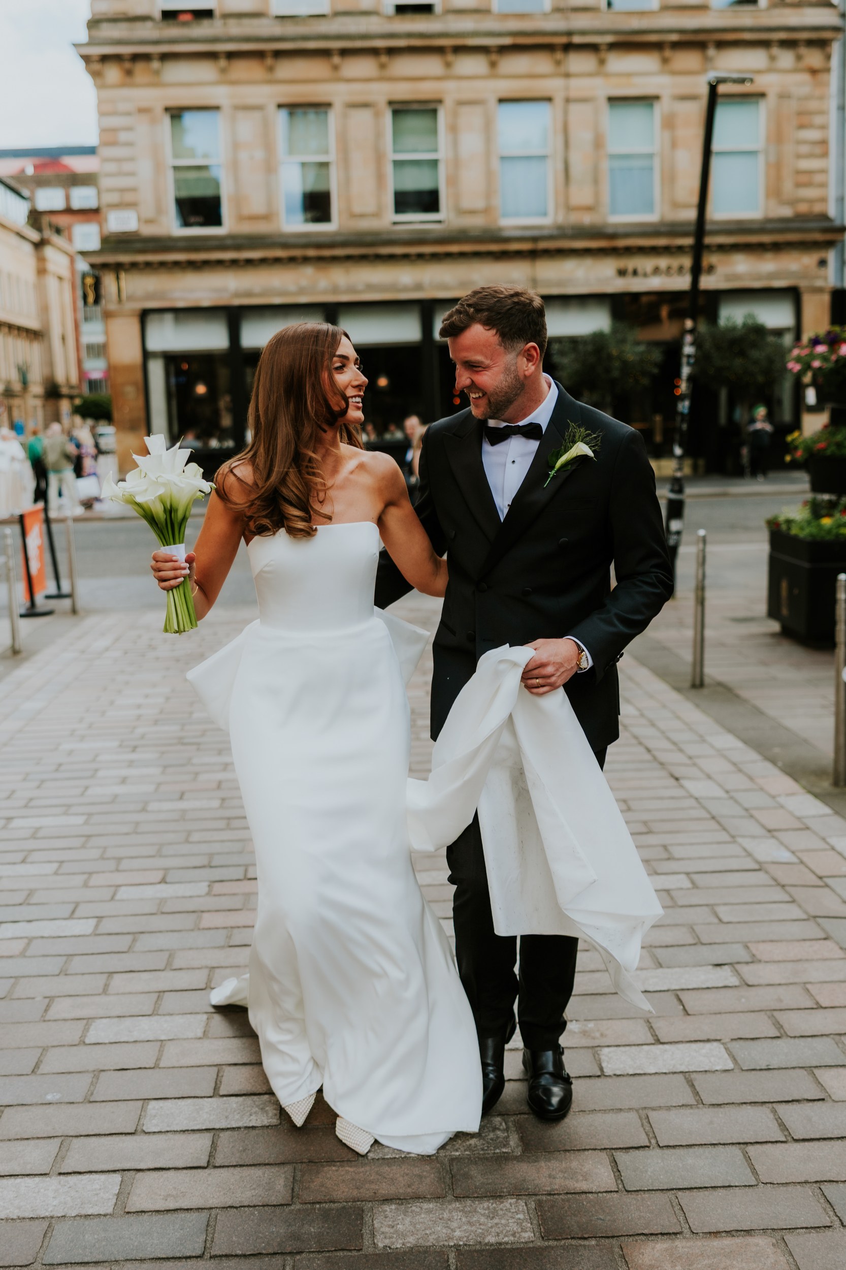 Citation Glasgow wedding photographer 47 Bride in strapless white gown holding calla lilies walks arm-in-arm with groom in a black tuxedo on a city street.