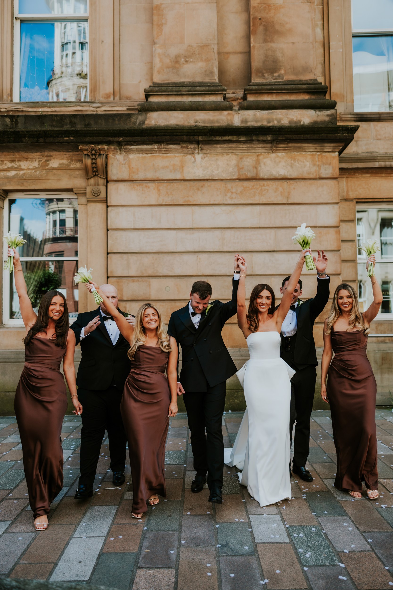 Citation Glasgow wedding photographer 46 Bride in white and groom with bridal party outside a stone building, raising bouquets; confetti on the pavement.