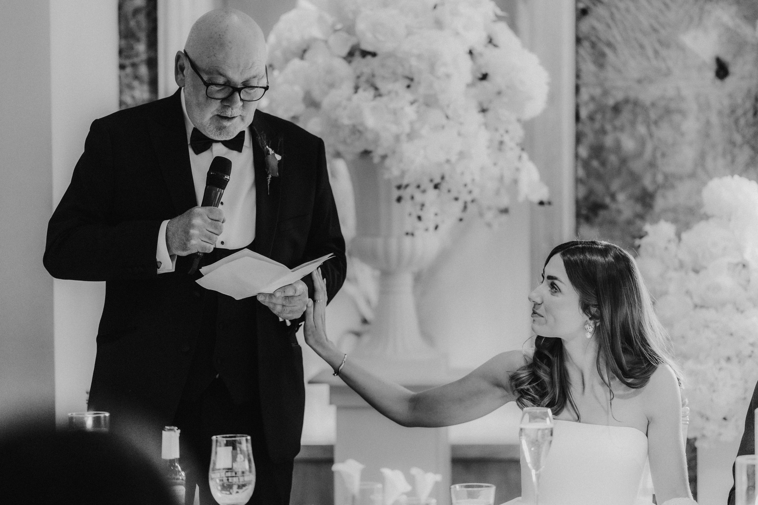 Citation Glasgow wedding photographer 74 Bride reaches toward a man in a tuxedo delivering a speech at a wedding reception with large floral arrangements behind them.