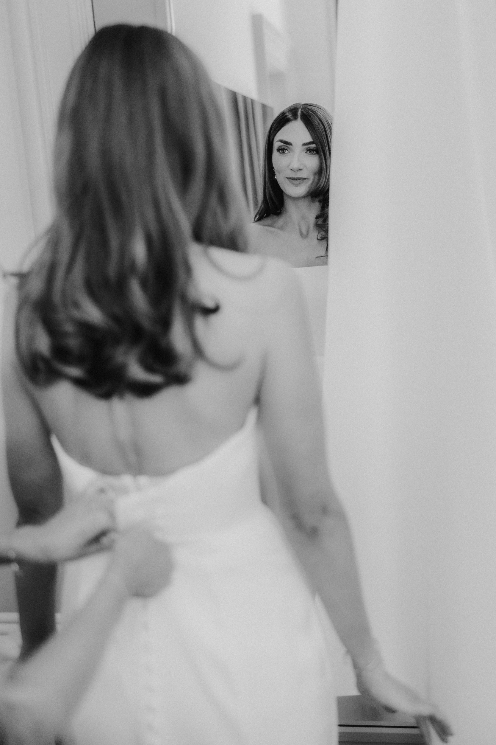 Citation Glasgow wedding photographer 13 Bride seen from behind looking at her reflection while someone fastens the buttons of her wedding dress.