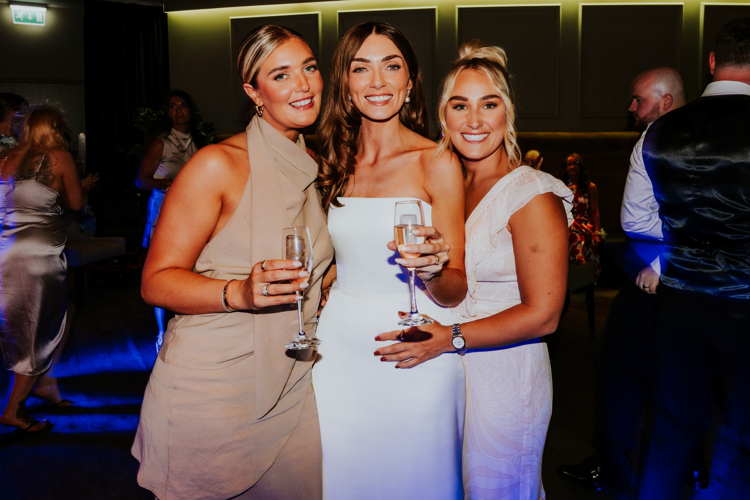 Citation Glasgow wedding photographer 87 Bride with two friends smiling and holding champagne flutes at an indoor wedding reception.