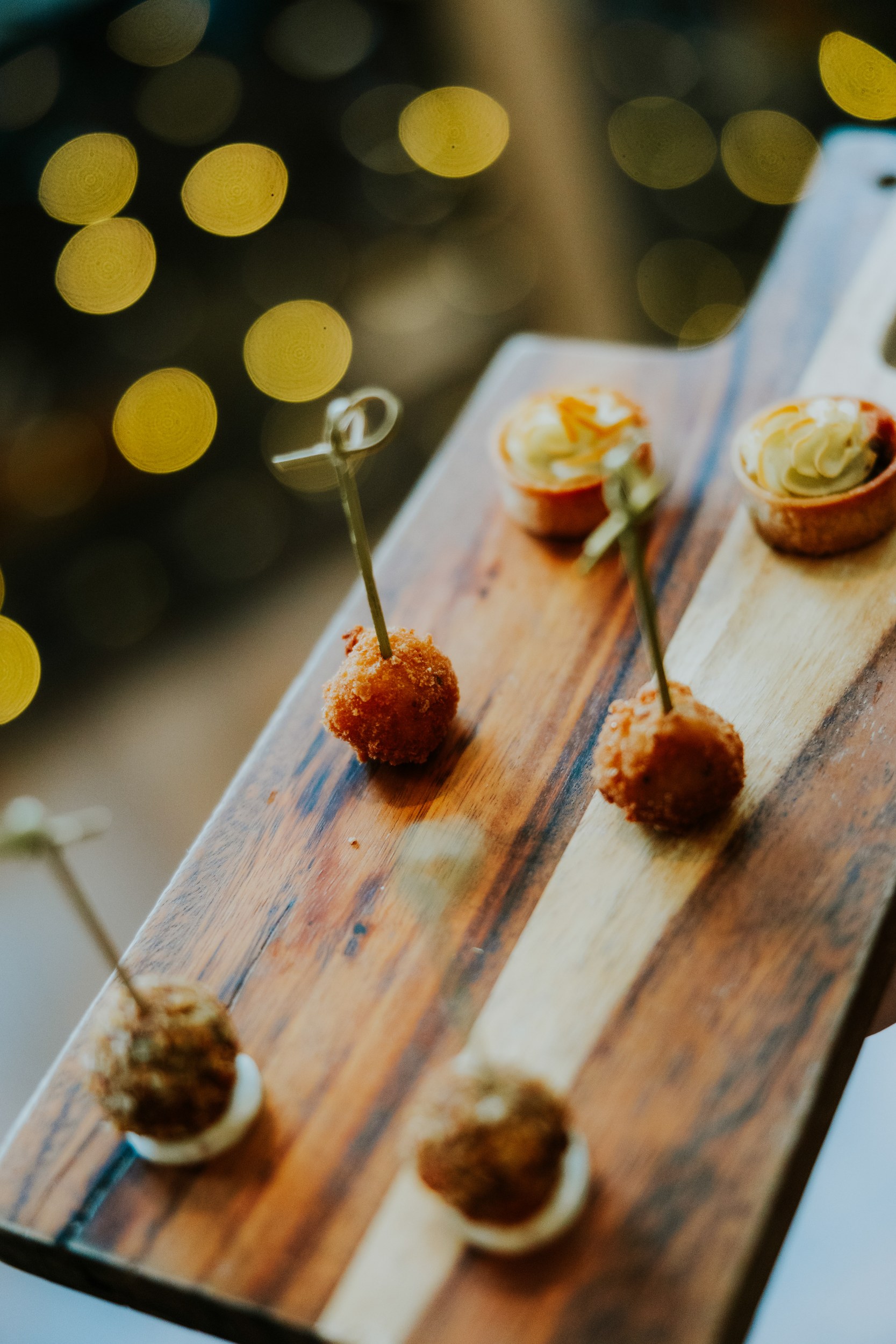 Citation Glasgow wedding photographer 69 Close-up of canapé skewers and mini tartlets on a wooden board with warm bokeh lights behind.