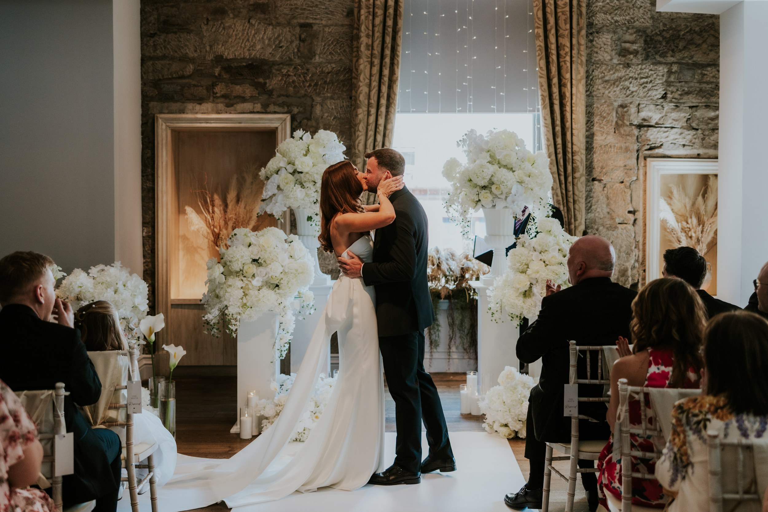 Citation Glasgow wedding photographer 34 Colette and Dean kiss at an indoor ceremony framed by white flowers and seated guests.