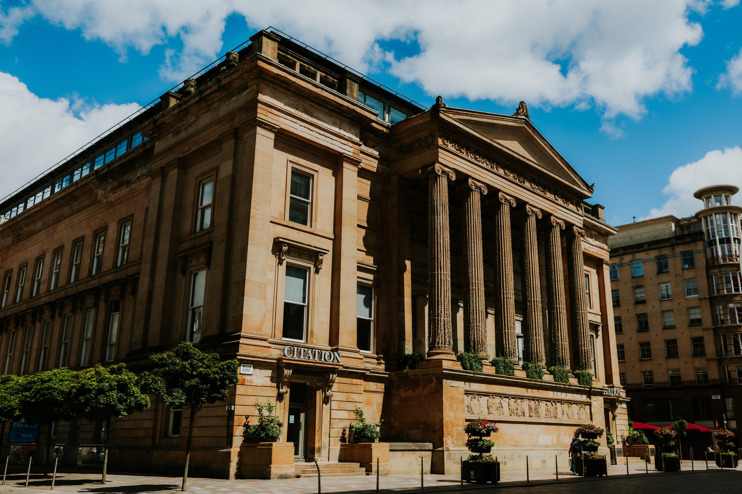 Citation Glasgow wedding photographer 1 Exterior of the Citation Glasgow building with neoclassical columns, entrance sign, trees and planters under a blue sky.