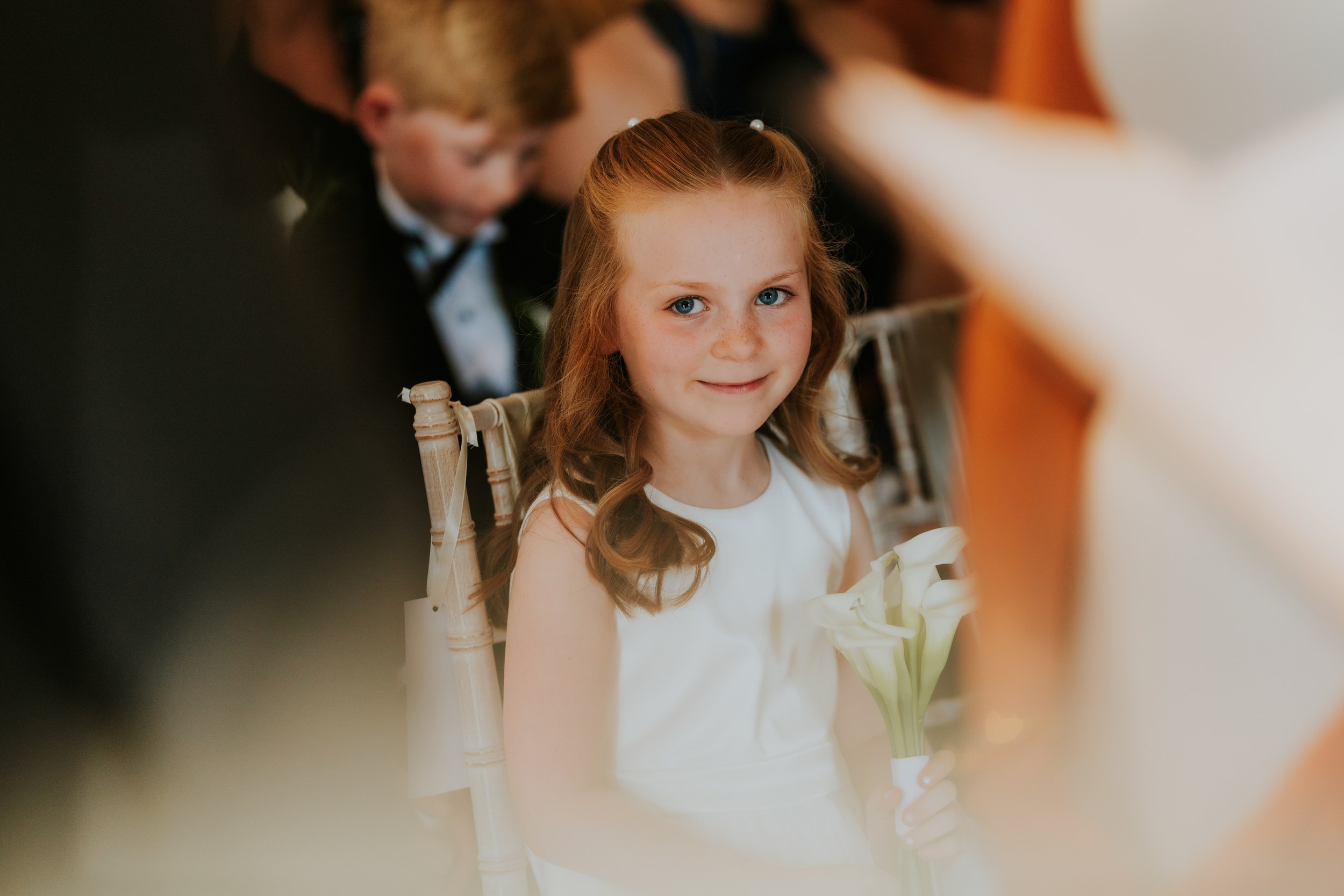 Citation Glasgow wedding photographer 28 Flower girl in white dress holding calla lilies, seated on a wooden chair and smiling with a blurred pageboy behind her.