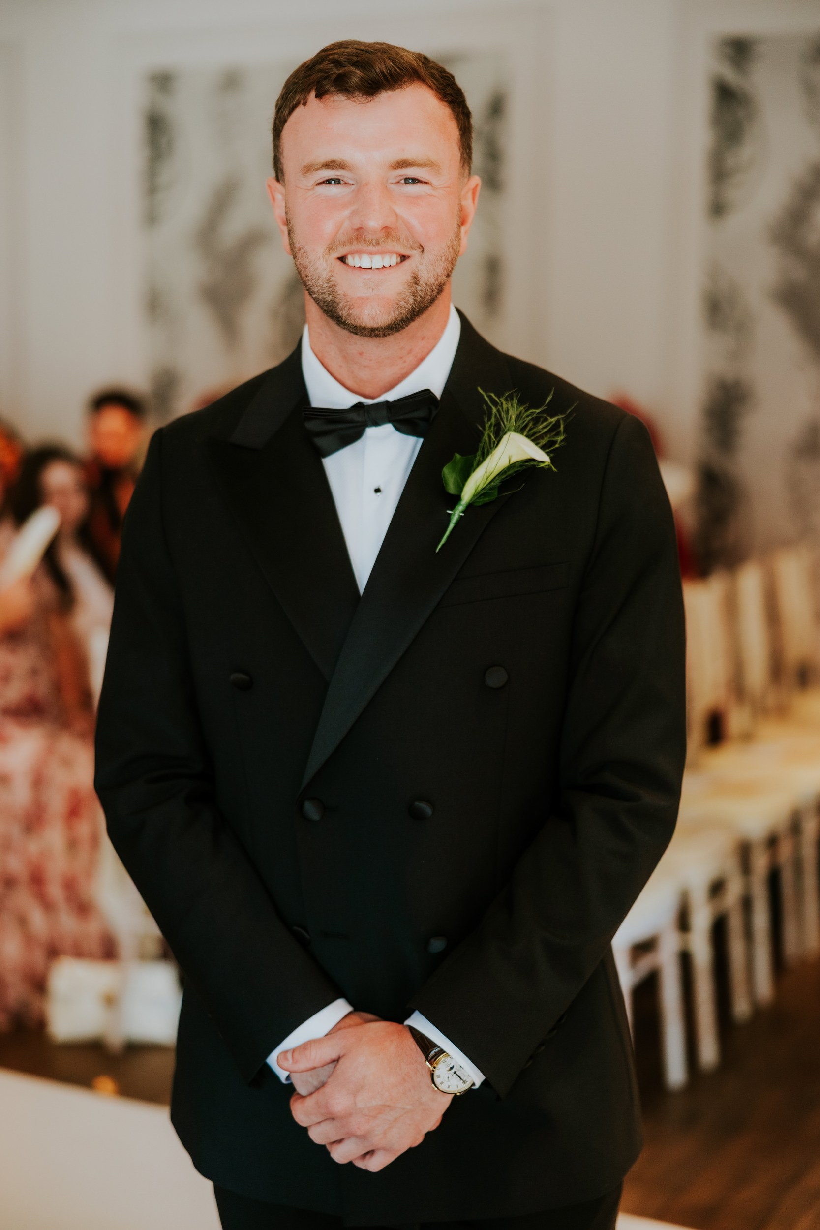 Citation Glasgow wedding photographer 19 Groom Dean smiling in black tuxedo with calla-lily boutonnière, hands clasped at an indoor wedding ceremony.