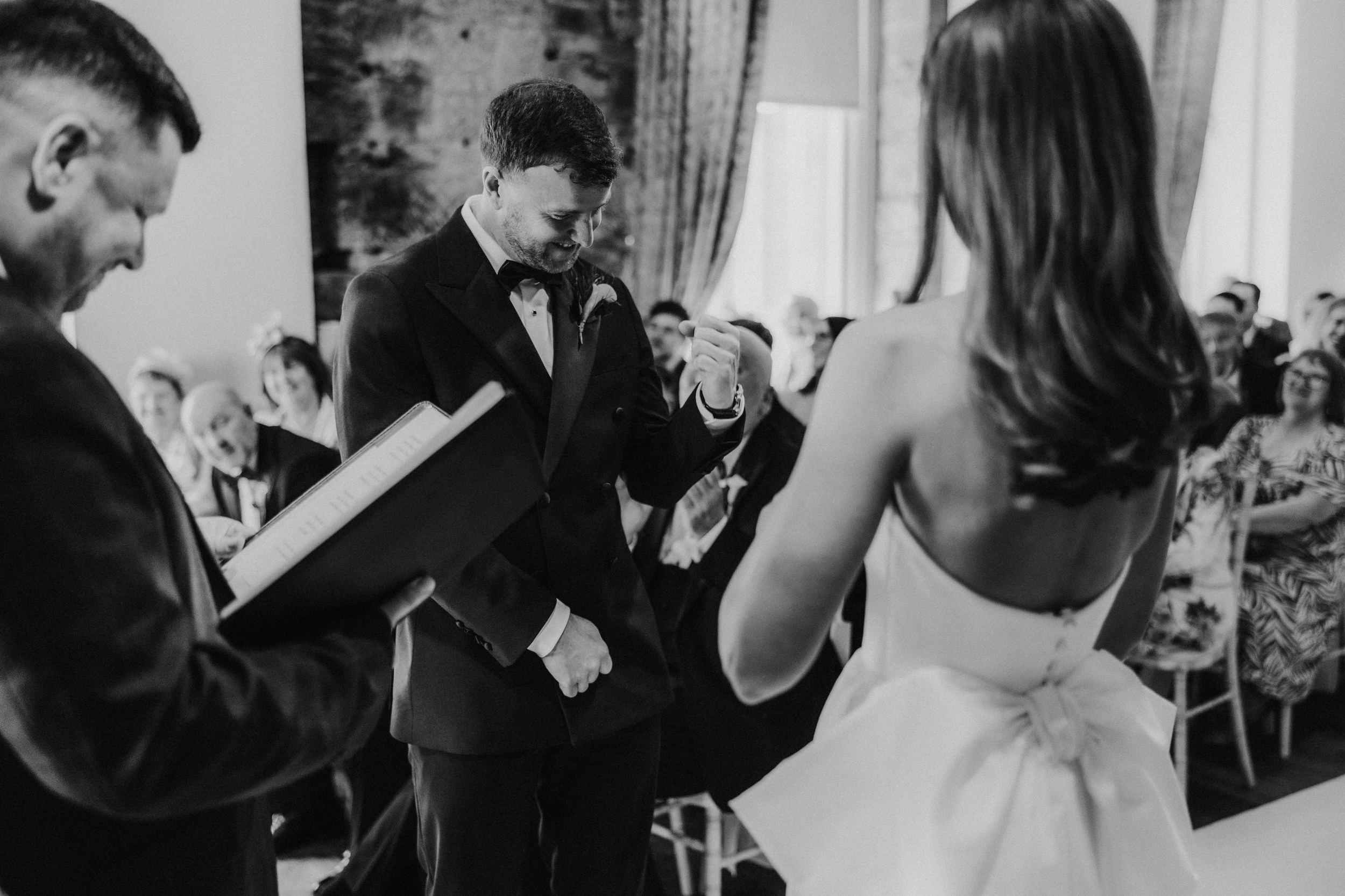 Citation Glasgow wedding photographer 29 Groom smiling and gesturing at his bride during the ceremony, officiant with a book and guests seated behind.