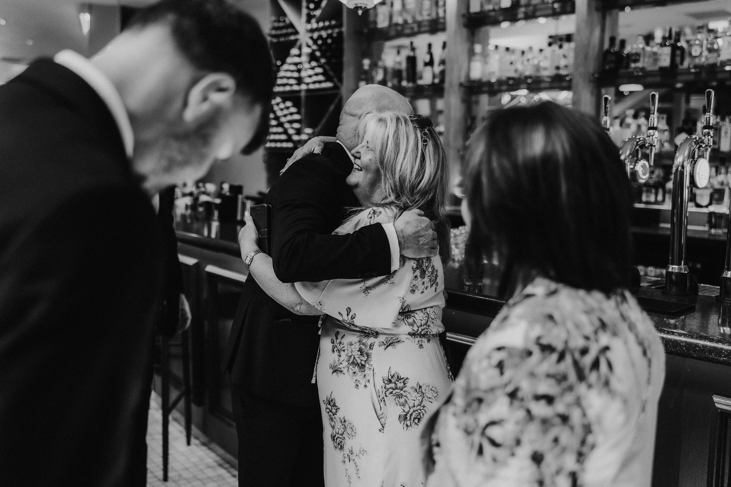 Citation Glasgow wedding photographer 8 Guests embrace at a venue bar, a woman smiling during the hug with bar taps and bottles behind them.