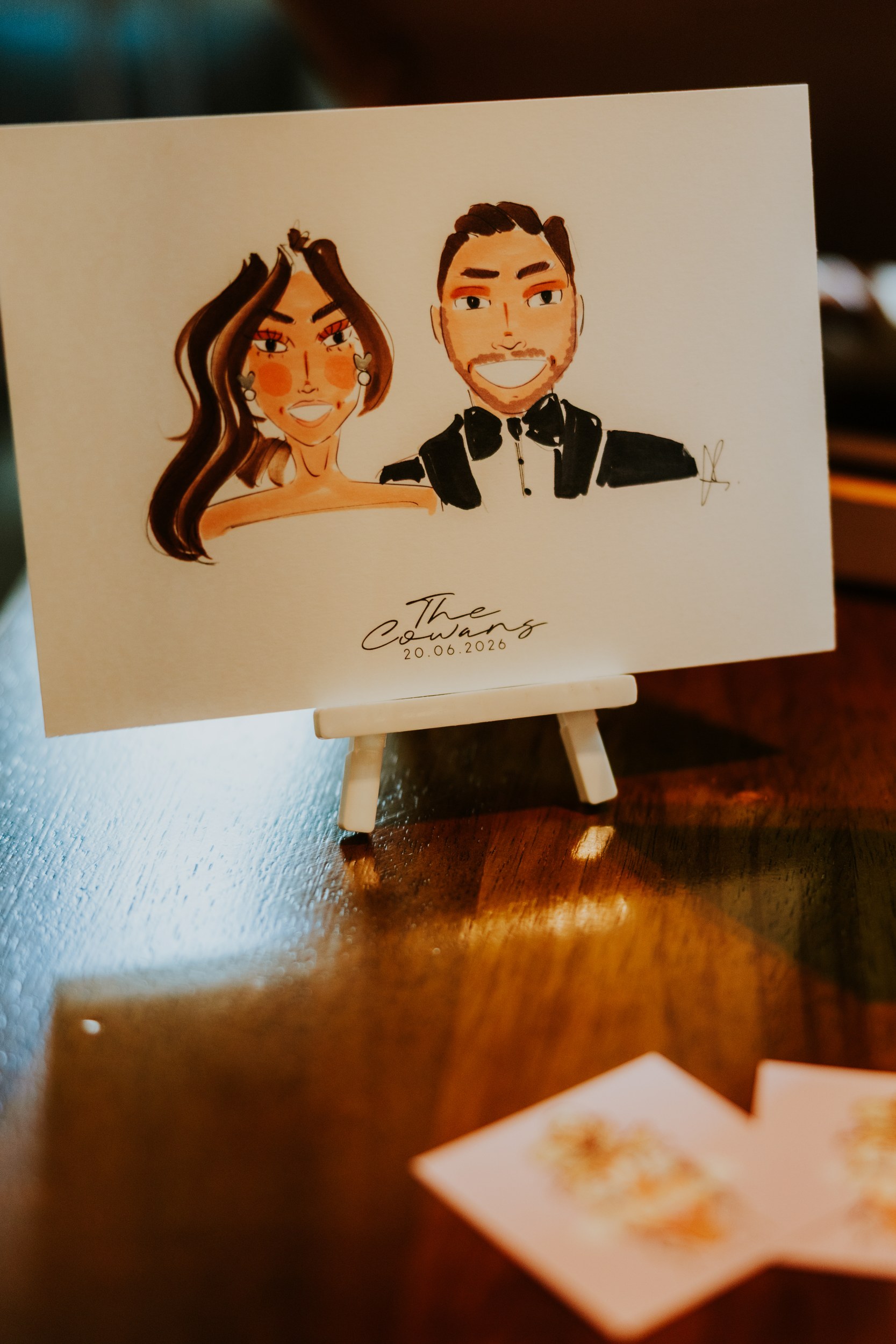 Citation Glasgow wedding photographer 63 Illustrated couple portrait on a small easel labelled 'The Cowans 20.06.2026' on a wooden table.