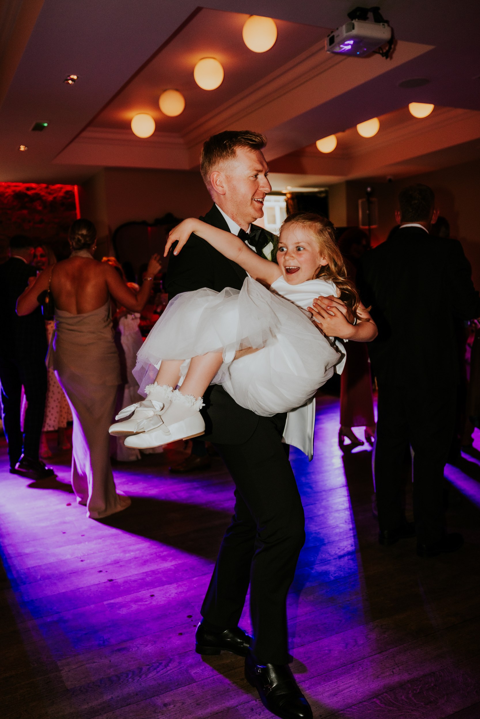 Citation Glasgow wedding photographer 79 Man in a suit carries a smiling young girl in a white dress on the wedding dancefloor.