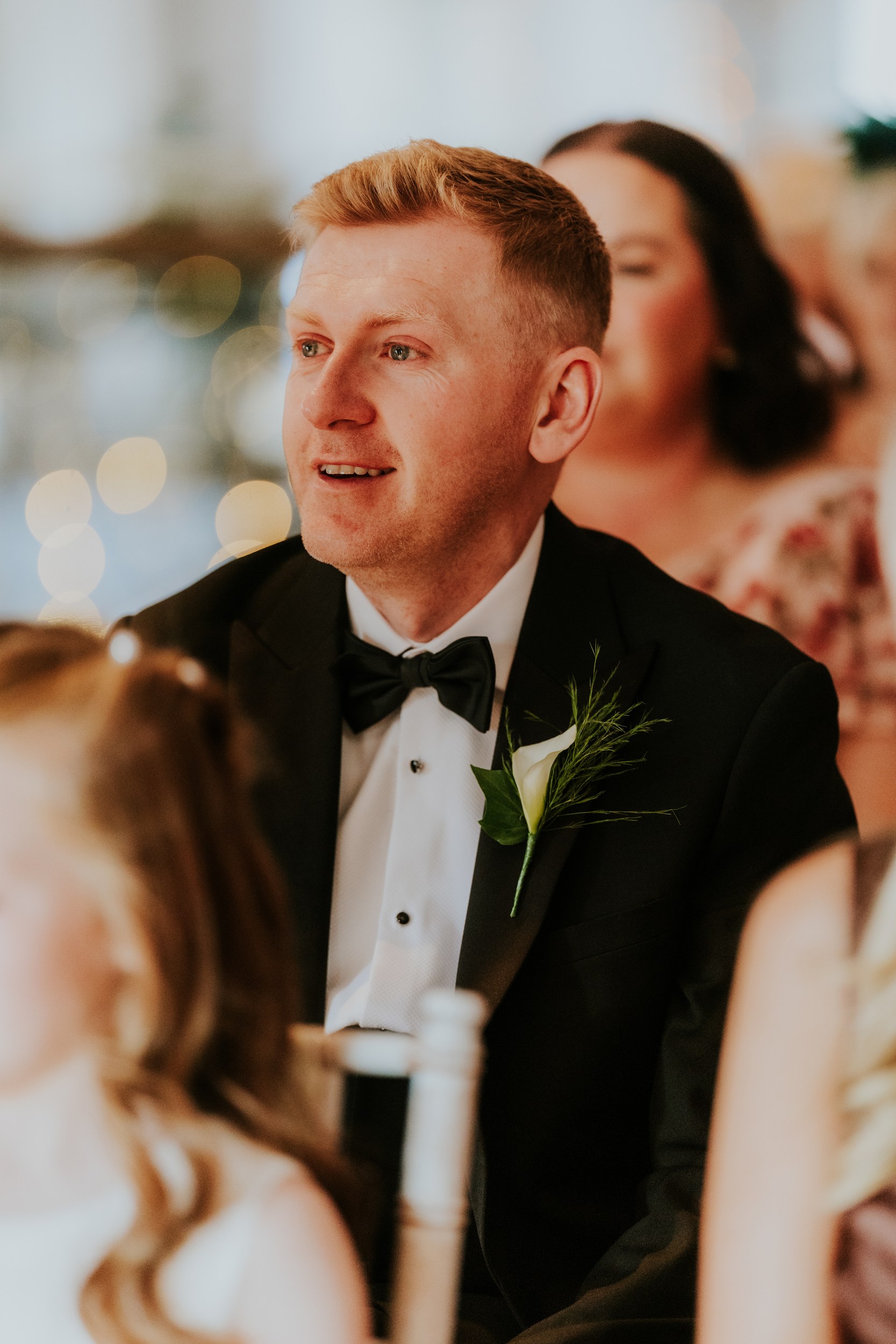 Citation Glasgow wedding photographer 31 Man in black tuxedo and bow tie with white calla lily boutonniere, seated at a reception with blurred guests and bokeh lights.