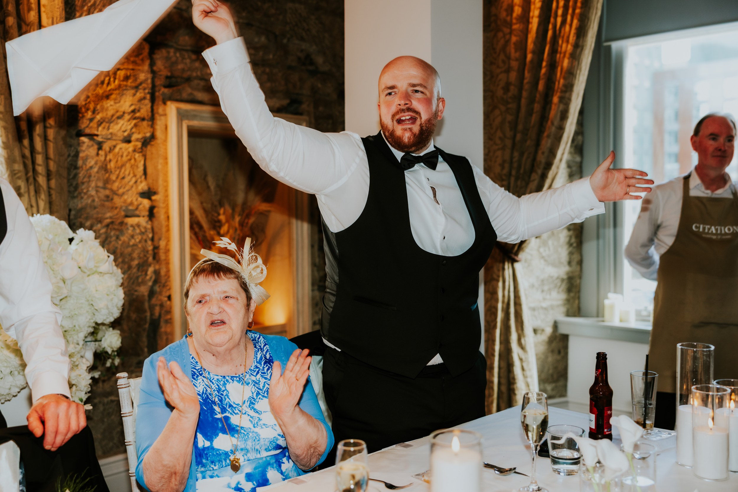 Citation Glasgow wedding photographer 73 Man in waistcoat and bow tie raising his arms at a wedding reception table, older woman clapping and staff wearing a Citation apron visible.