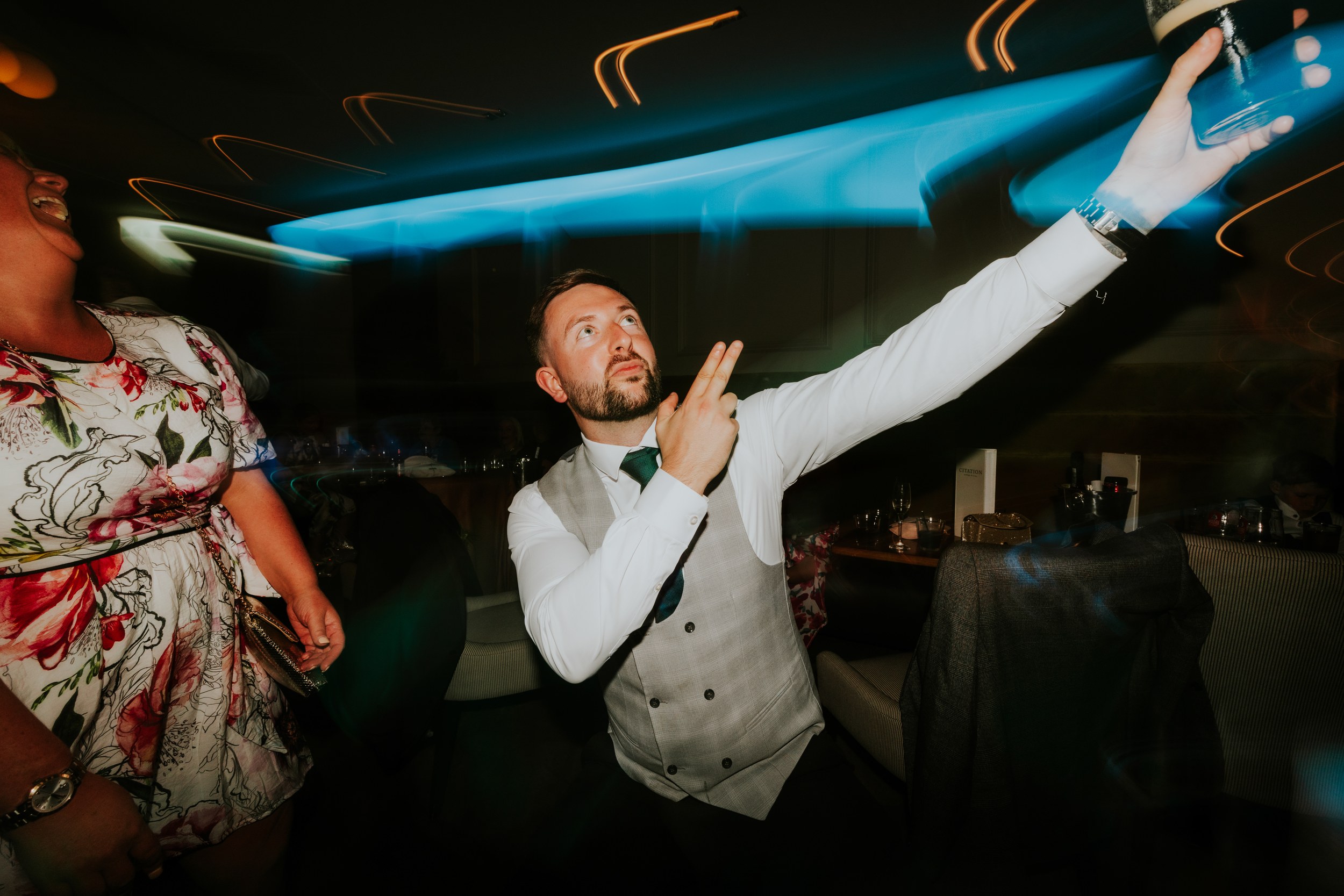 Citation Glasgow wedding photographer 90 Man in waistcoat raises a drink and points amid motion-blurred blue lights at a reception, woman in a floral dress at left.