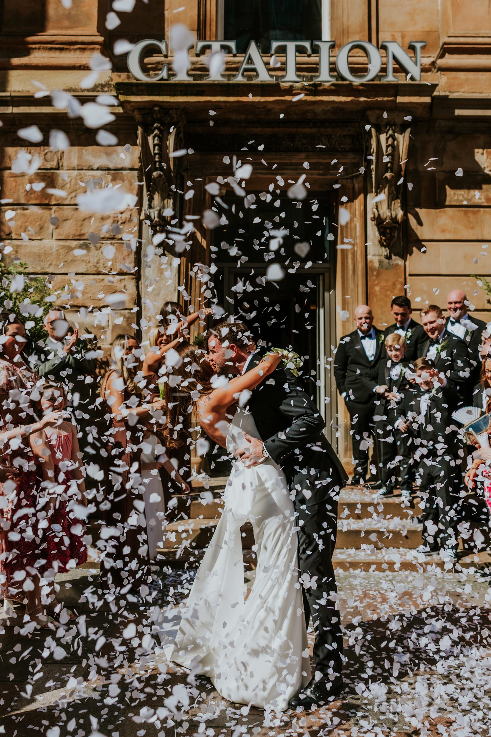 Citation Glasgow wedding photographer 40 Newlyweds kiss under a shower of confetti outside the Citation Glasgow entrance with wedding party watching.