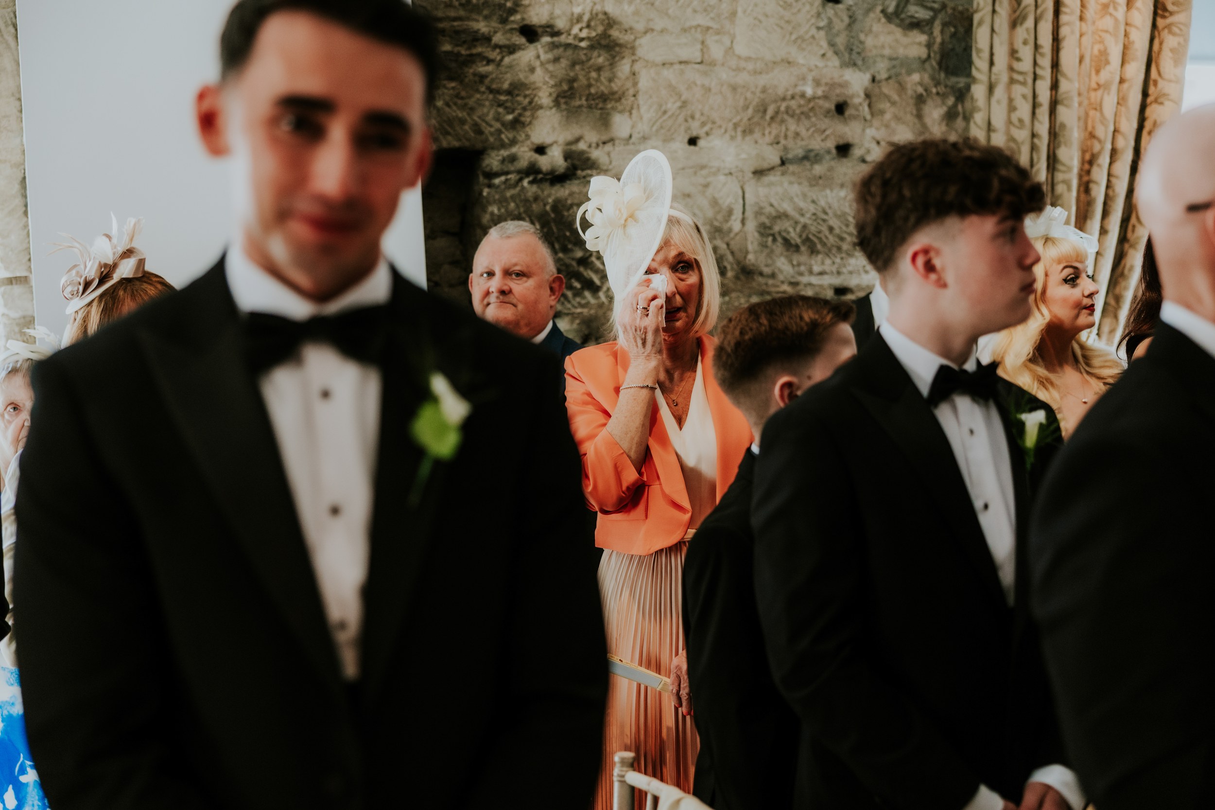 Citation Glasgow wedding photographer 26 Older woman in a cream hat wipes a tear among guests in black tuxedos at an indoor wedding ceremony.