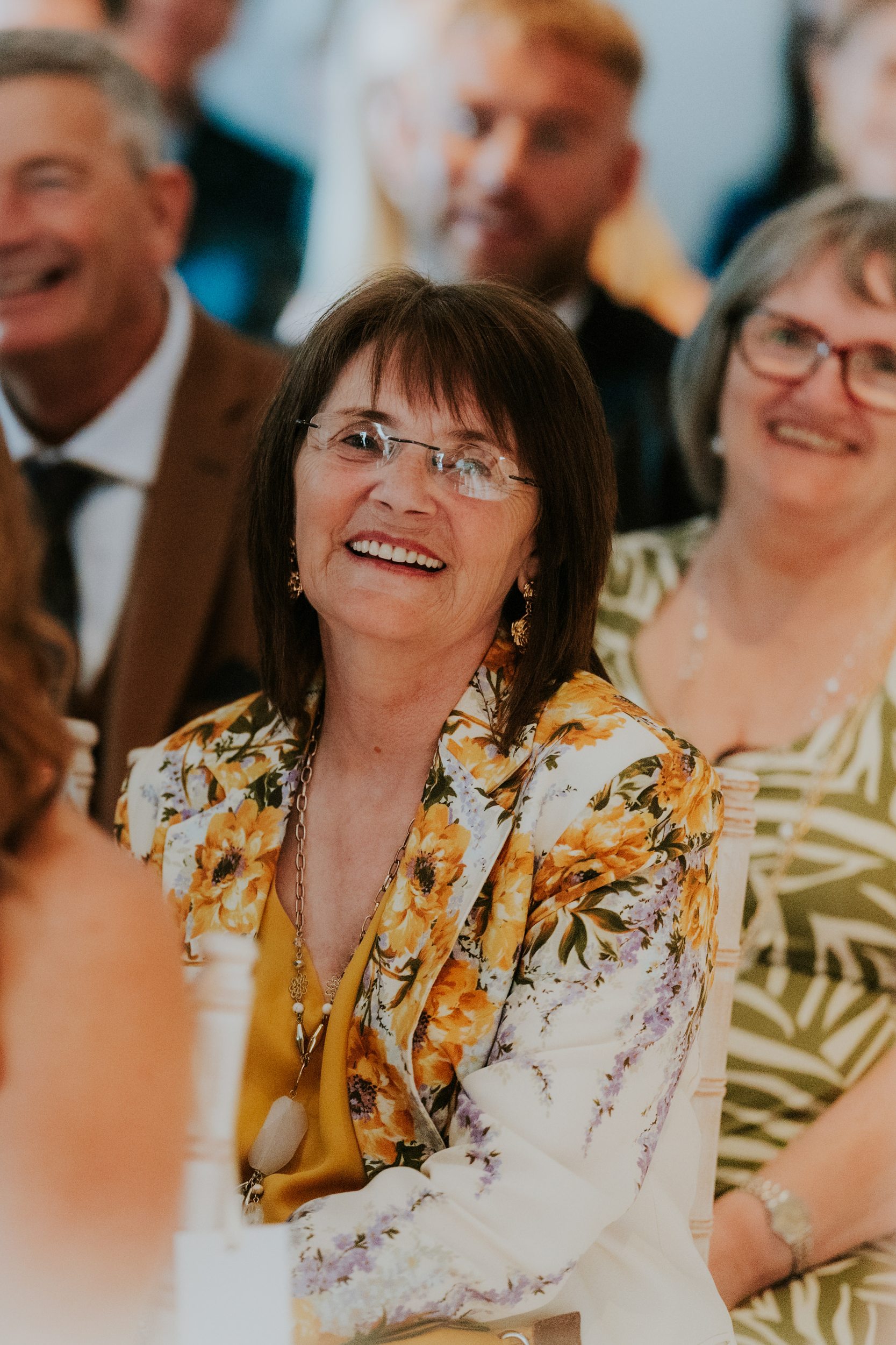 Citation Glasgow wedding photographer 32 Smiling female wedding guest in glasses wearing a yellow floral jacket, seated among other guests.
