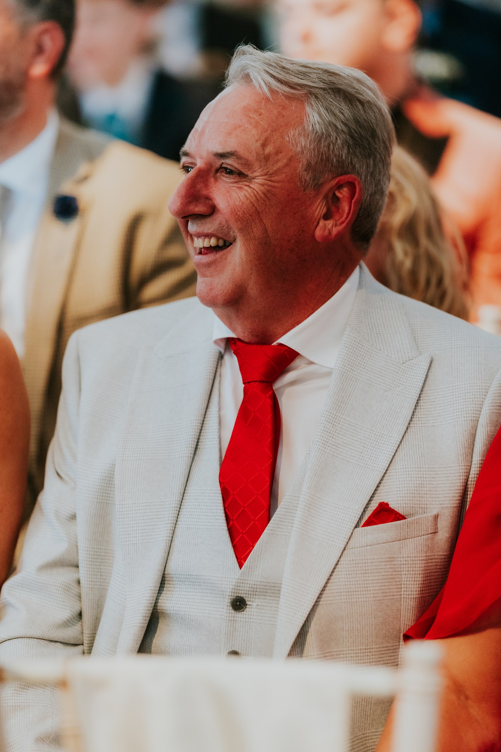 Citation Glasgow wedding photographer 30 Smiling man in light grey suit with red tie and pocket square at a wedding reception.