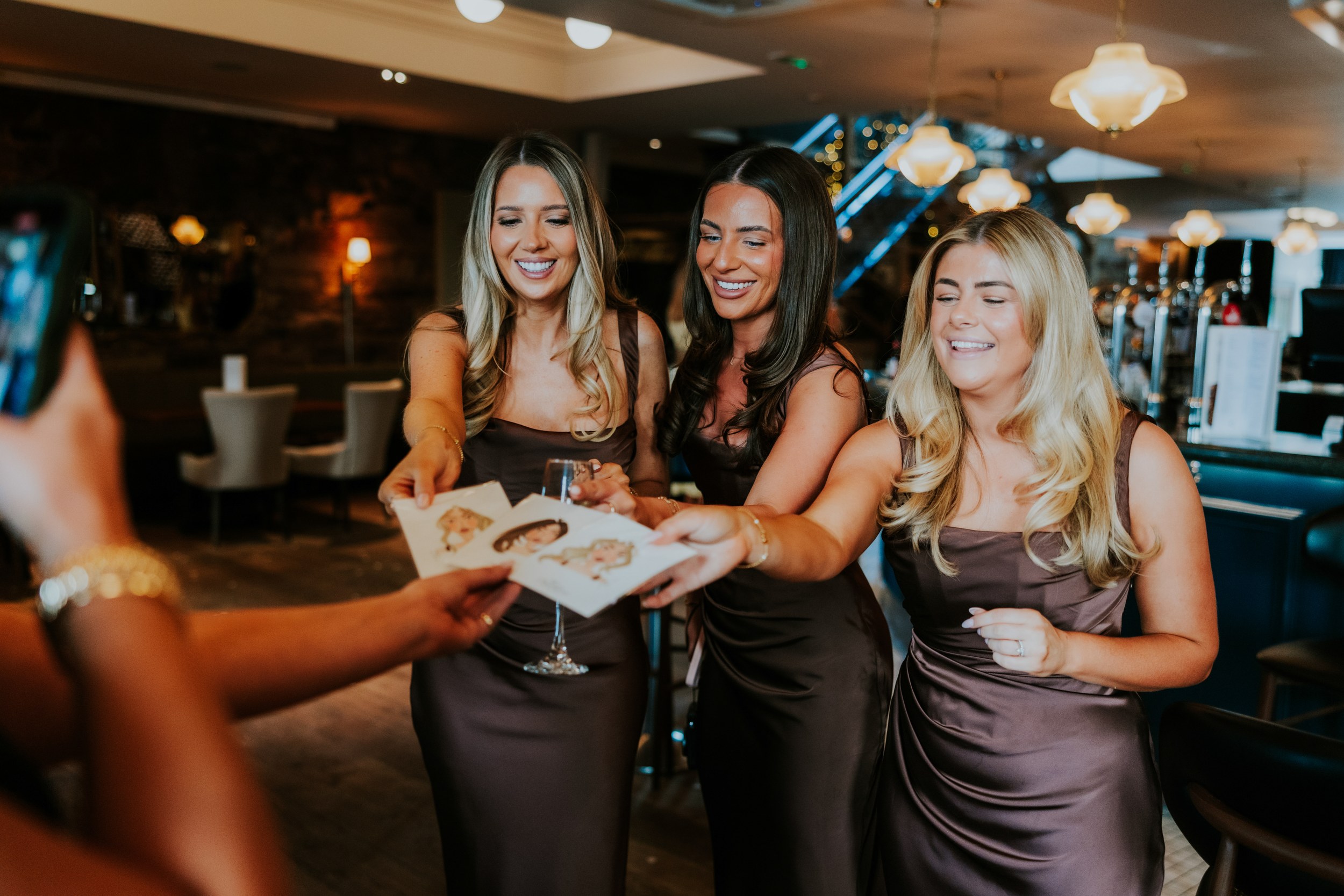 Citation Glasgow wedding photographer 65 Three bridesmaids in brown satin dresses taking illustrated cards at a venue bar, smiling.