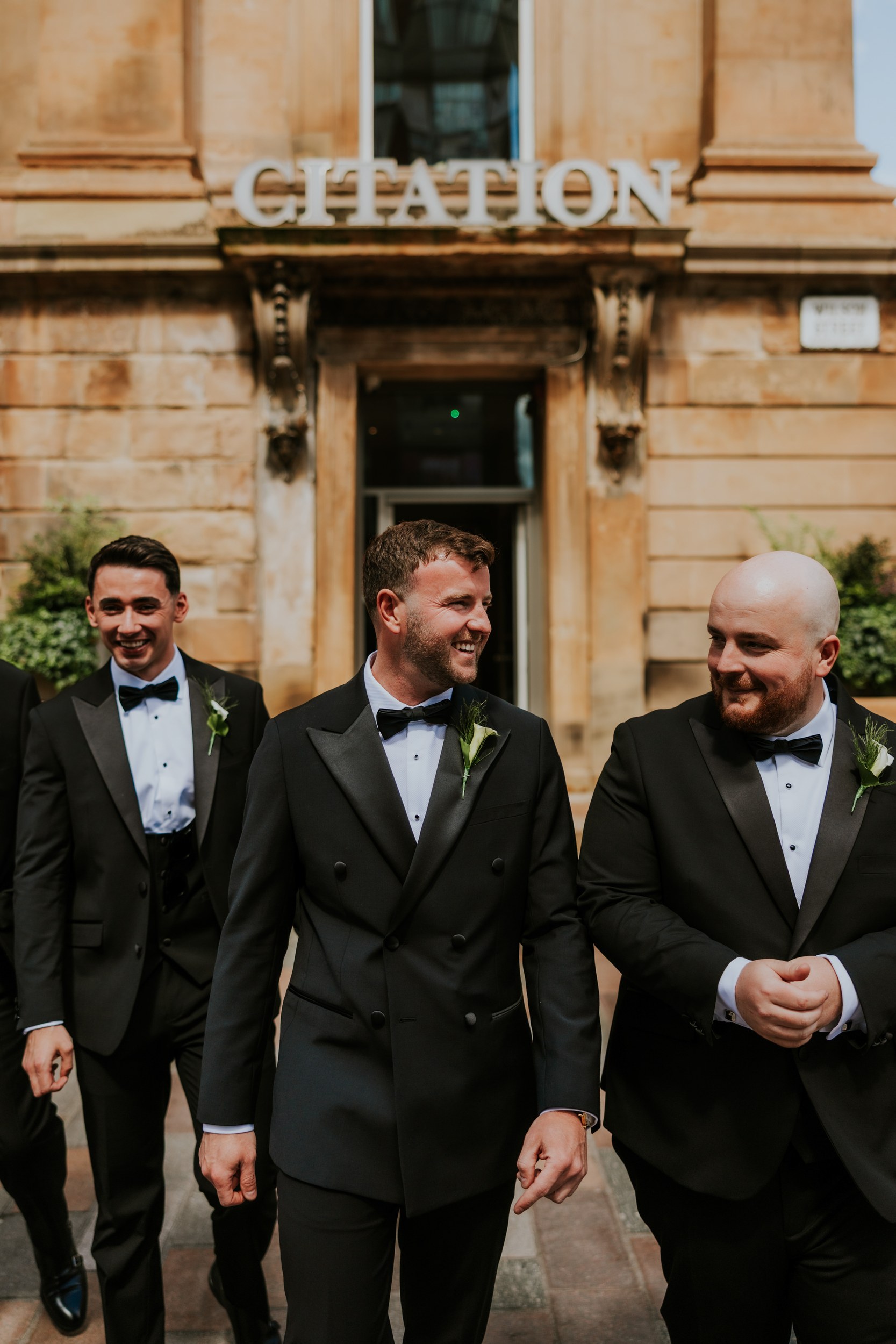 Citation Glasgow wedding photographer 10 Three groomsmen in black tuxedos smiling outside the Citation Glasgow entrance beneath the 'CITATION' sign.