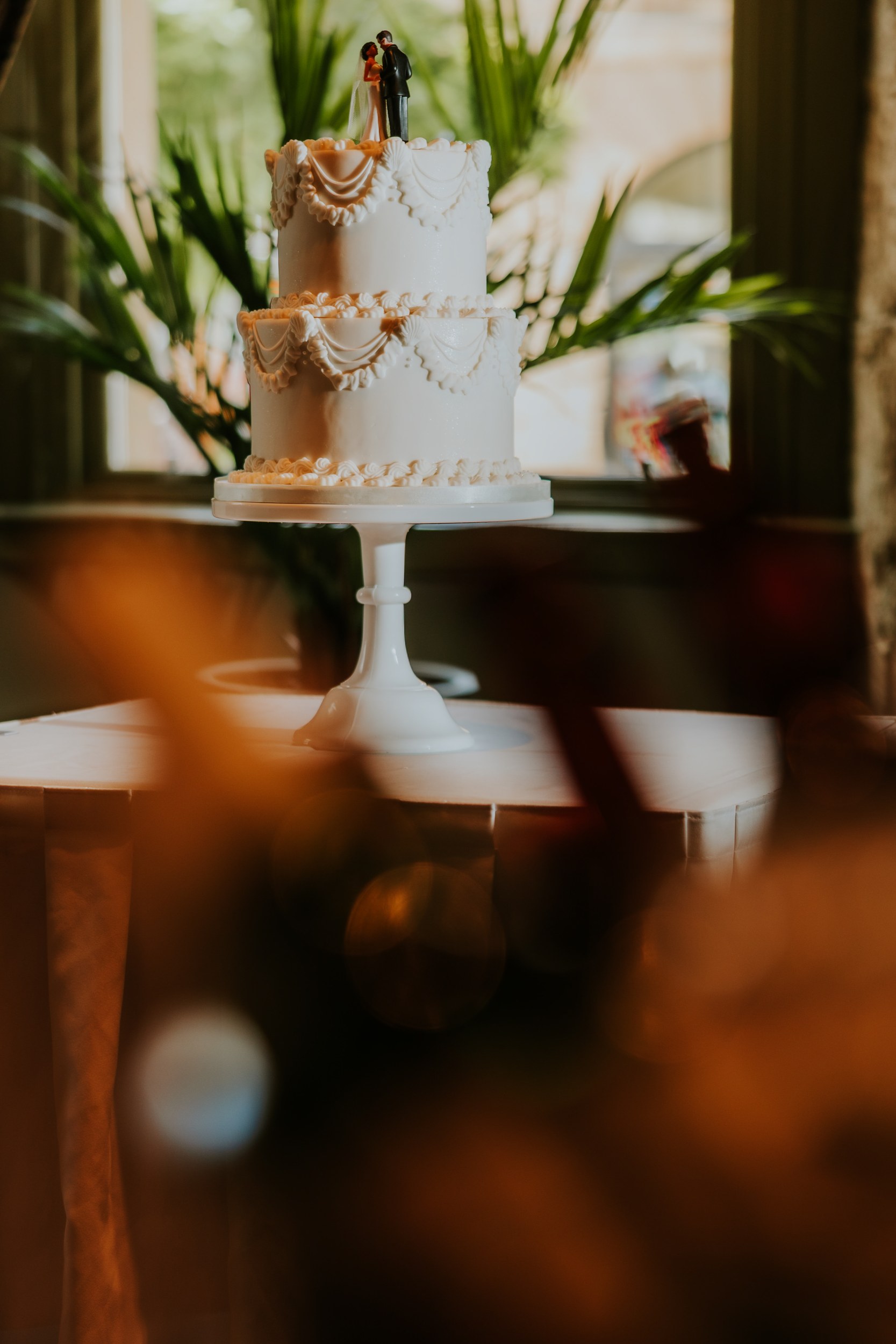 Citation Glasgow wedding photographer 4 Two-tier white wedding cake with bride-and-groom topper on a white stand, blurred plants behind.