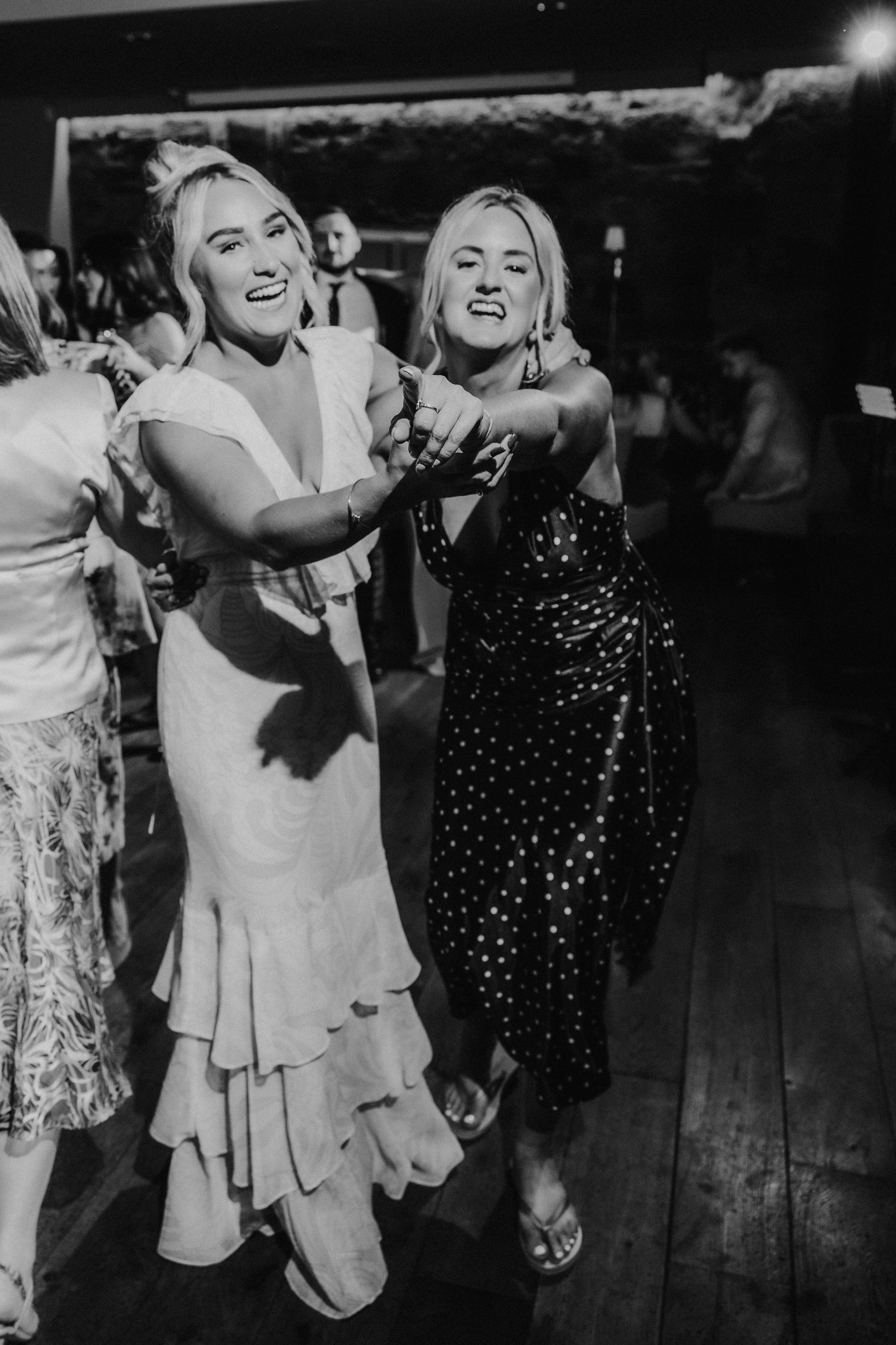 Citation Glasgow wedding photographer 86 Two women on a dance floor smiling and pointing at the camera, one in a light ruffled dress and one in a dark polka-dot dress.