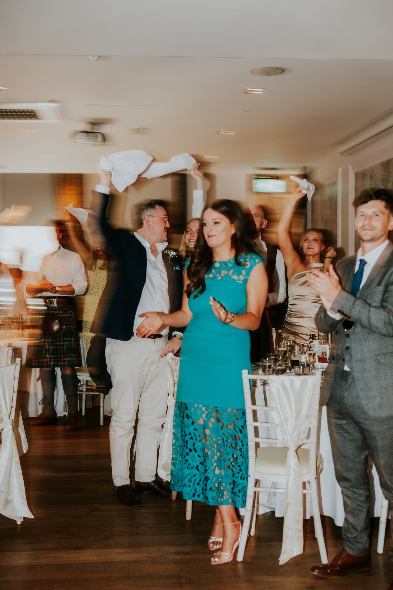 Citation Glasgow wedding photographer 71 Wedding guests cheering and waving napkins at an indoor reception.