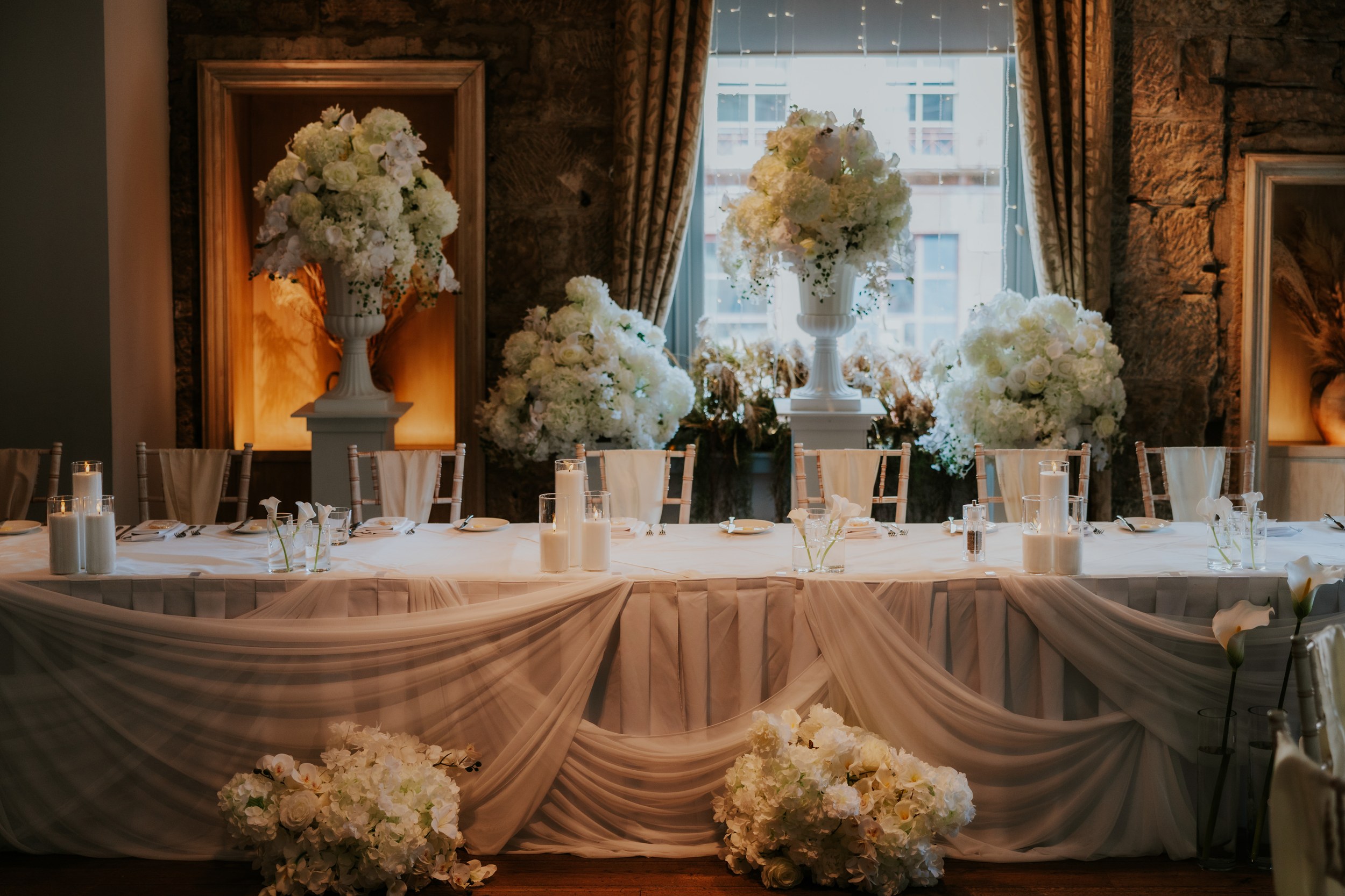 Citation Glasgow wedding photographer 60 Wedding head table with white floral urns, candles and draped fabric in a stone-walled room at Citation Glasgow.