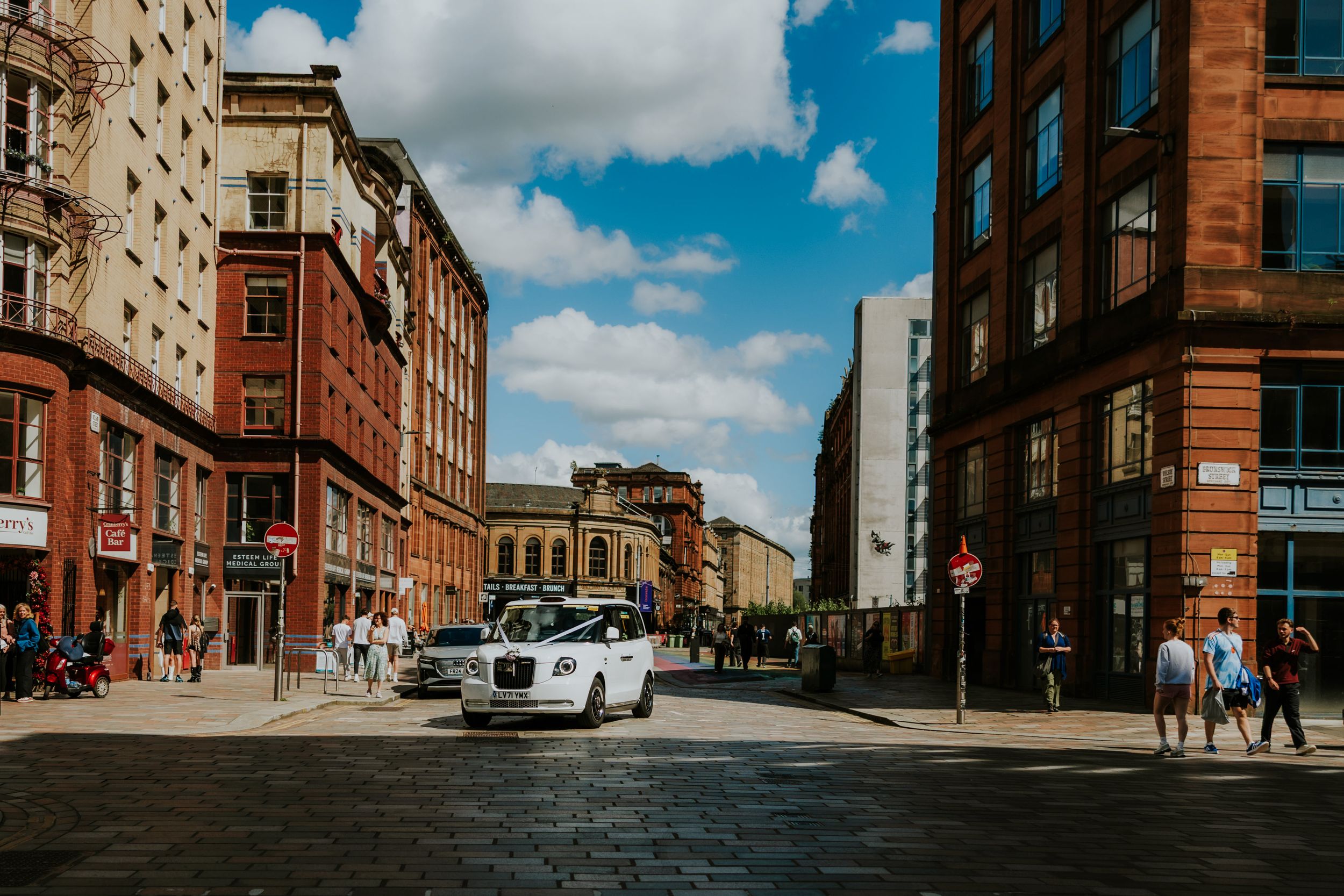 Citation Glasgow wedding photographer 18 White wedding taxi with ribbons on a Glasgow street between red sandstone buildings under a blue sky.