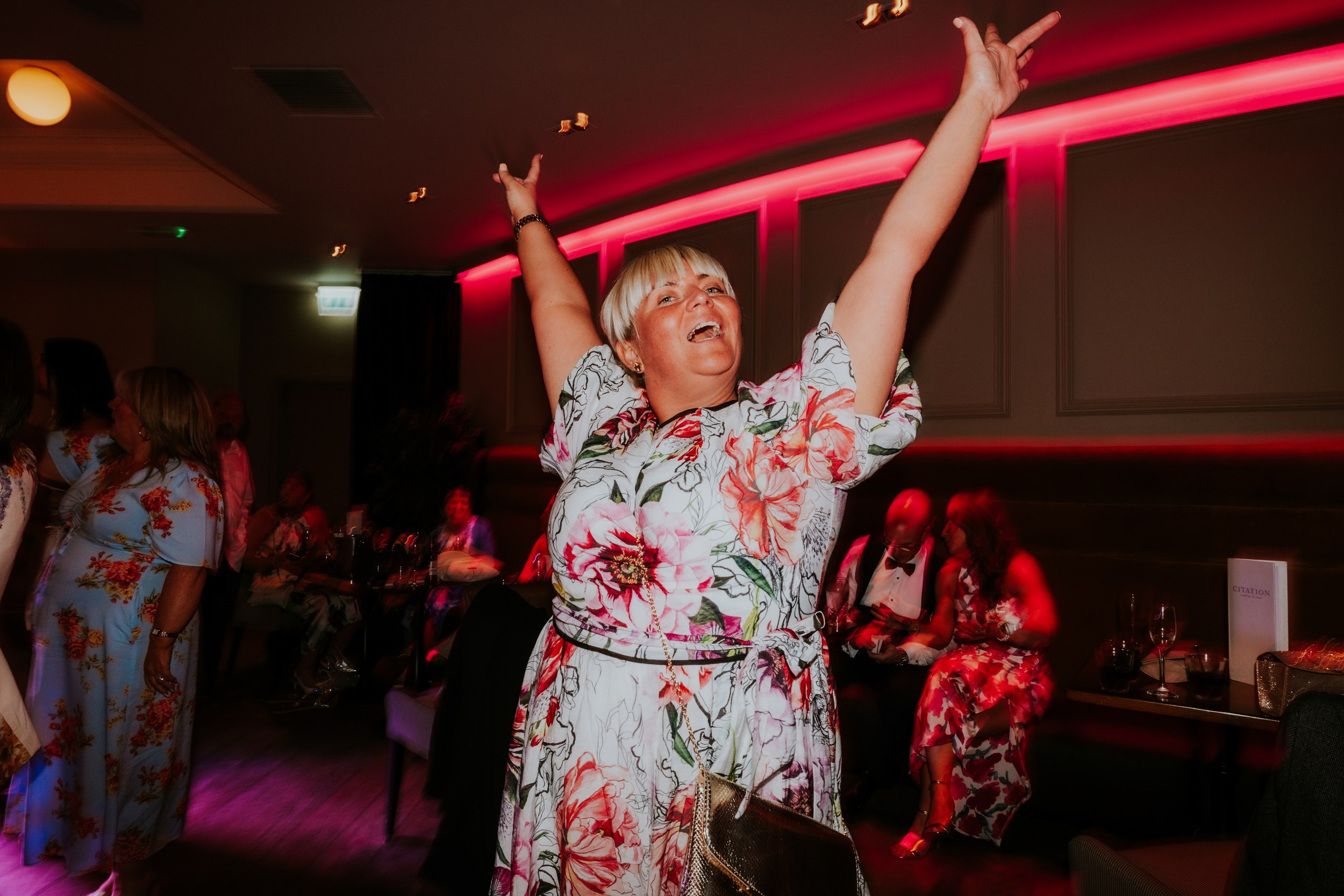 Citation Glasgow wedding photographer 89 Woman in floral dress dancing with arms raised under pink uplighting at a wedding reception.