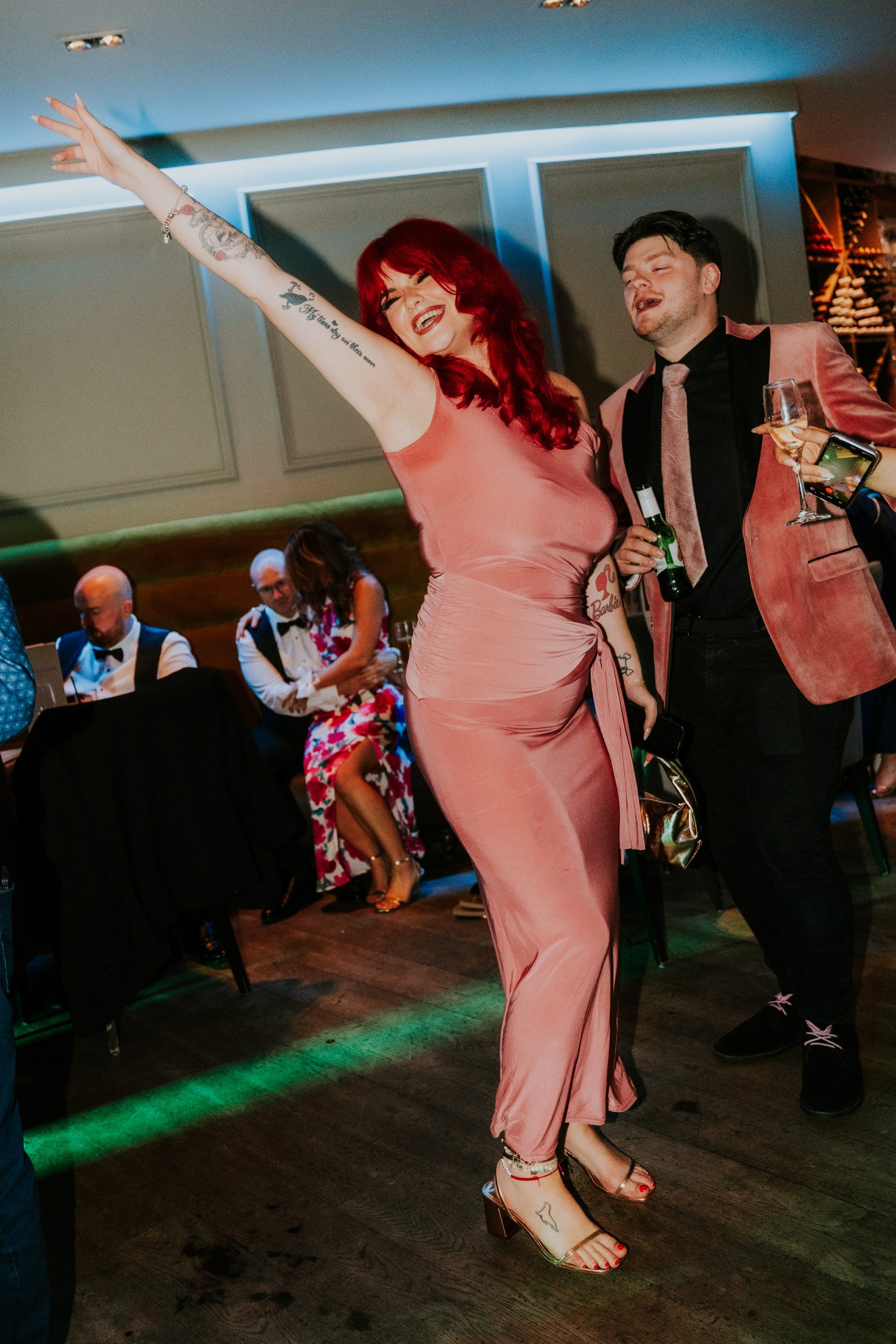 Citation Glasgow wedding photographer 84 Woman in pink dress dancing with raised arm beside a man in a pink jacket holding drinks at a wedding reception.