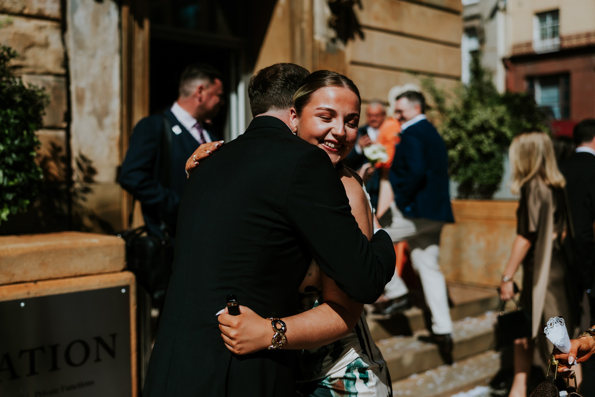 Citation Glasgow wedding photographer 41 Woman smiling with eyes closed as she hugs a person in a dark suit outside a stone wedding venue.