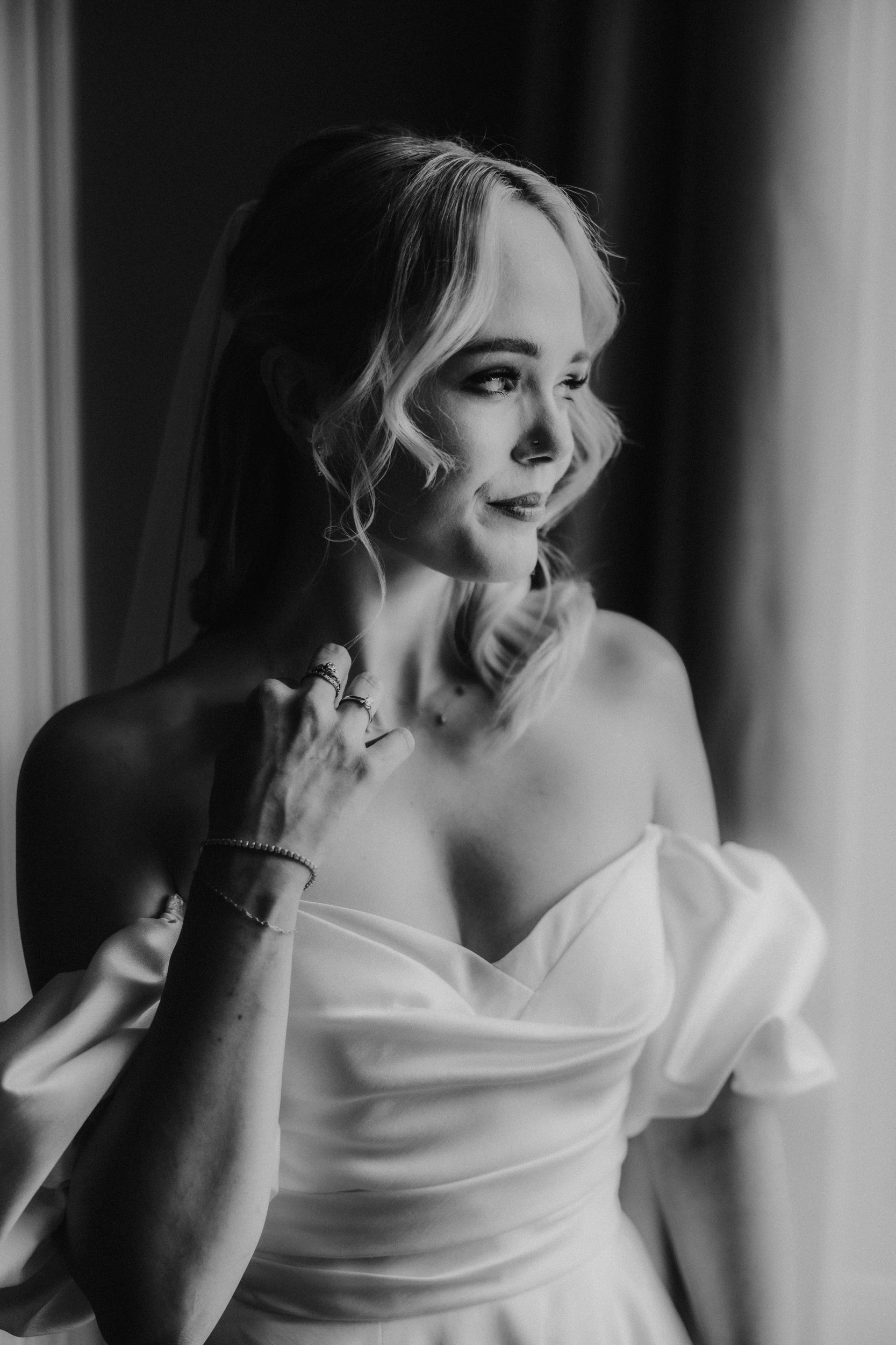 Black-and-white portrait of a bride in an off-the-shoulder dress looking toward a window, hand with rings at her chest.