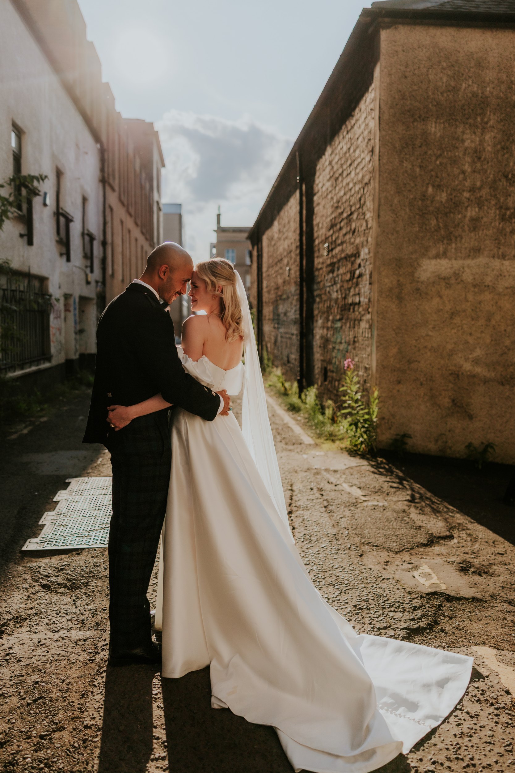 Bride and groom embracing in a sunlit alley, bride in white gown and veil and groom in dark jacket.