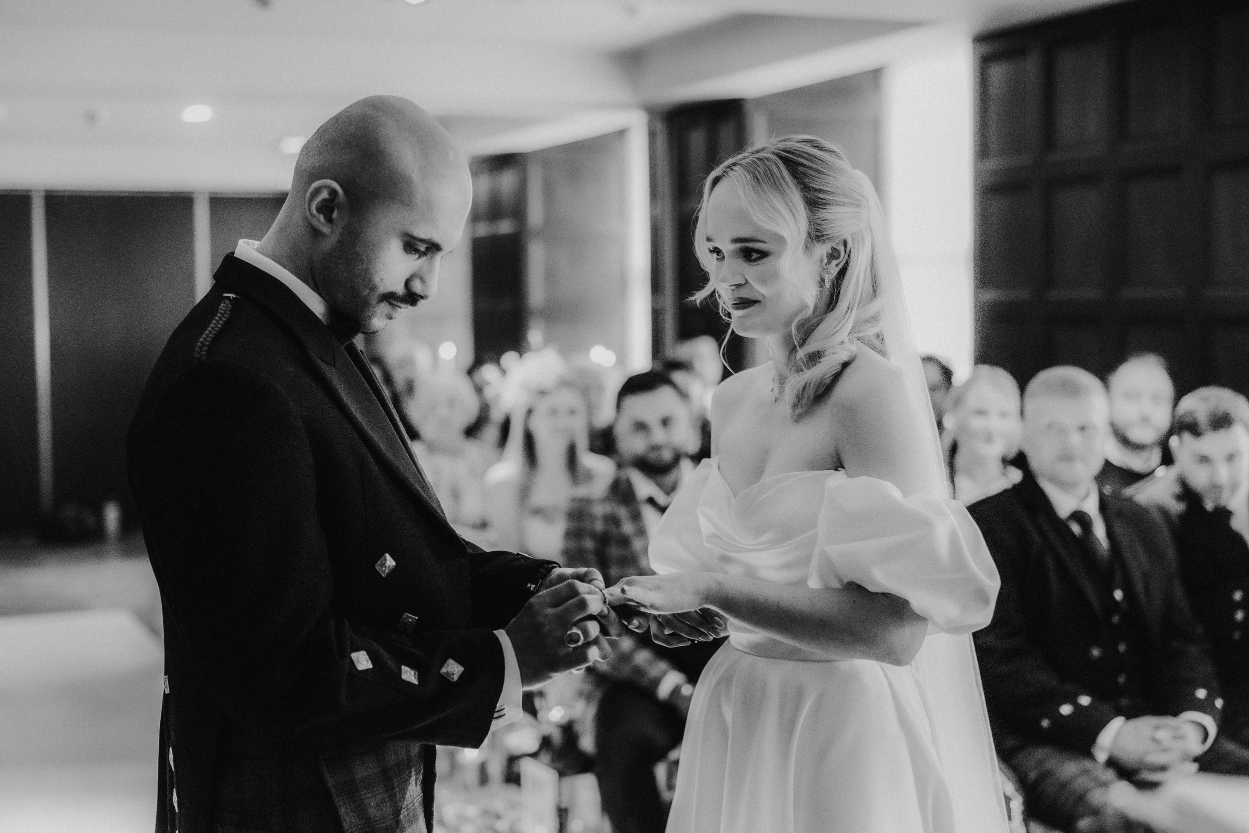 Bride and groom exchange rings at an indoor ceremony with seated guests behind them.