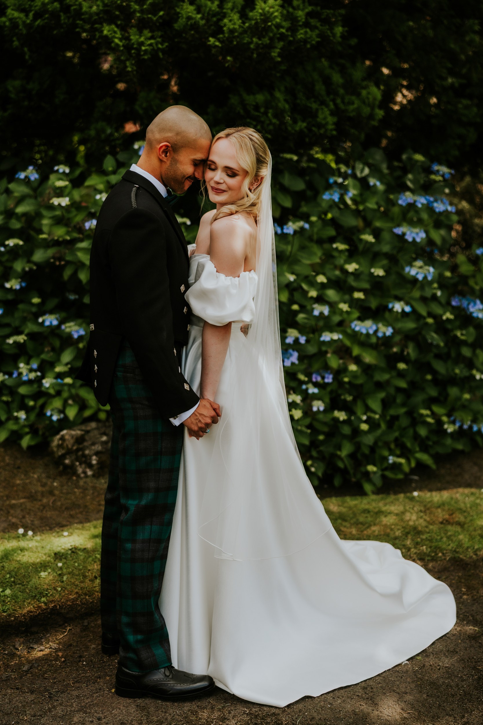 Bride and groom hold hands and touch foreheads in front of hydrangea bushes.