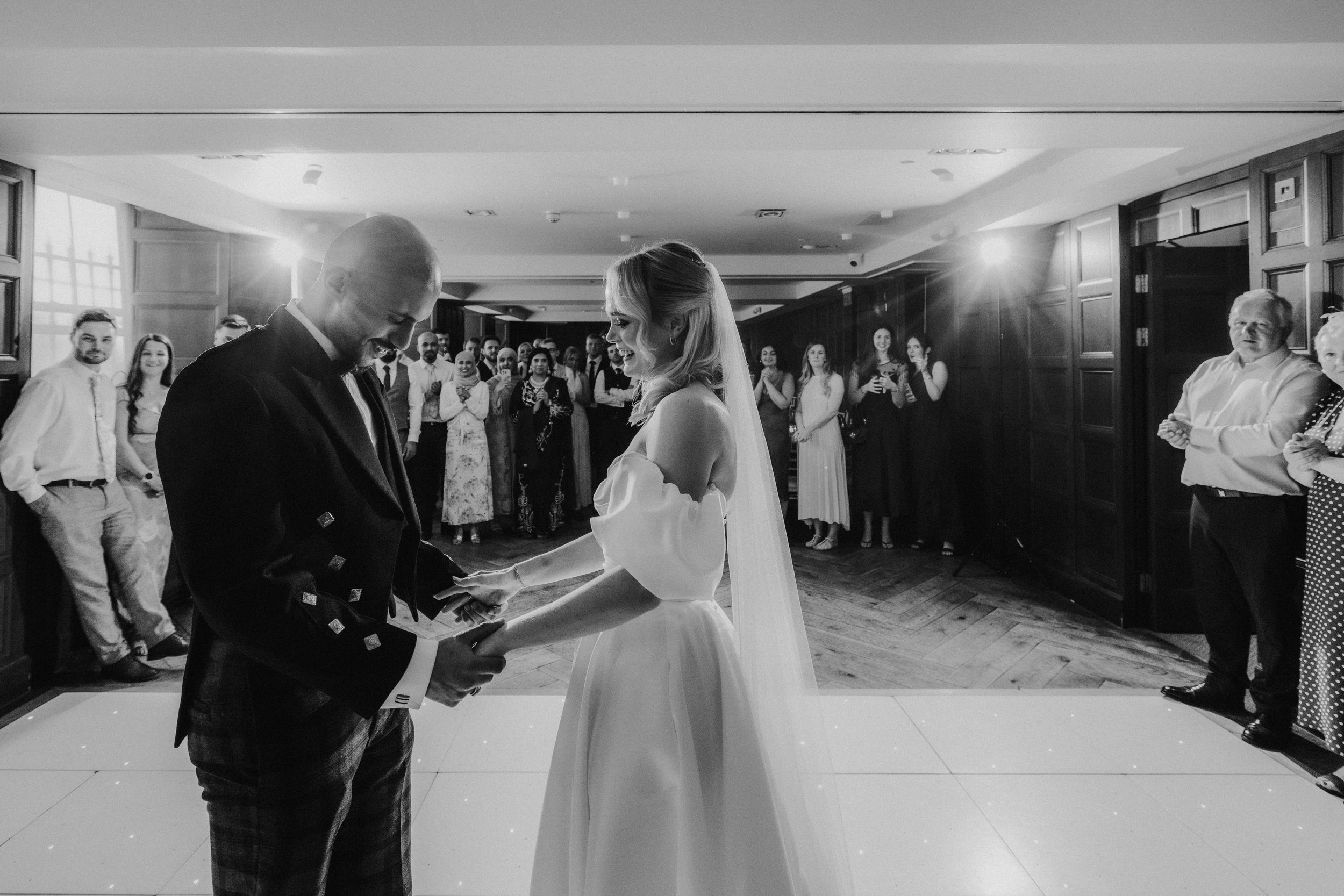 Bride and groom hold hands on a lit dancefloor as guests watch in a wood‑panelled room.