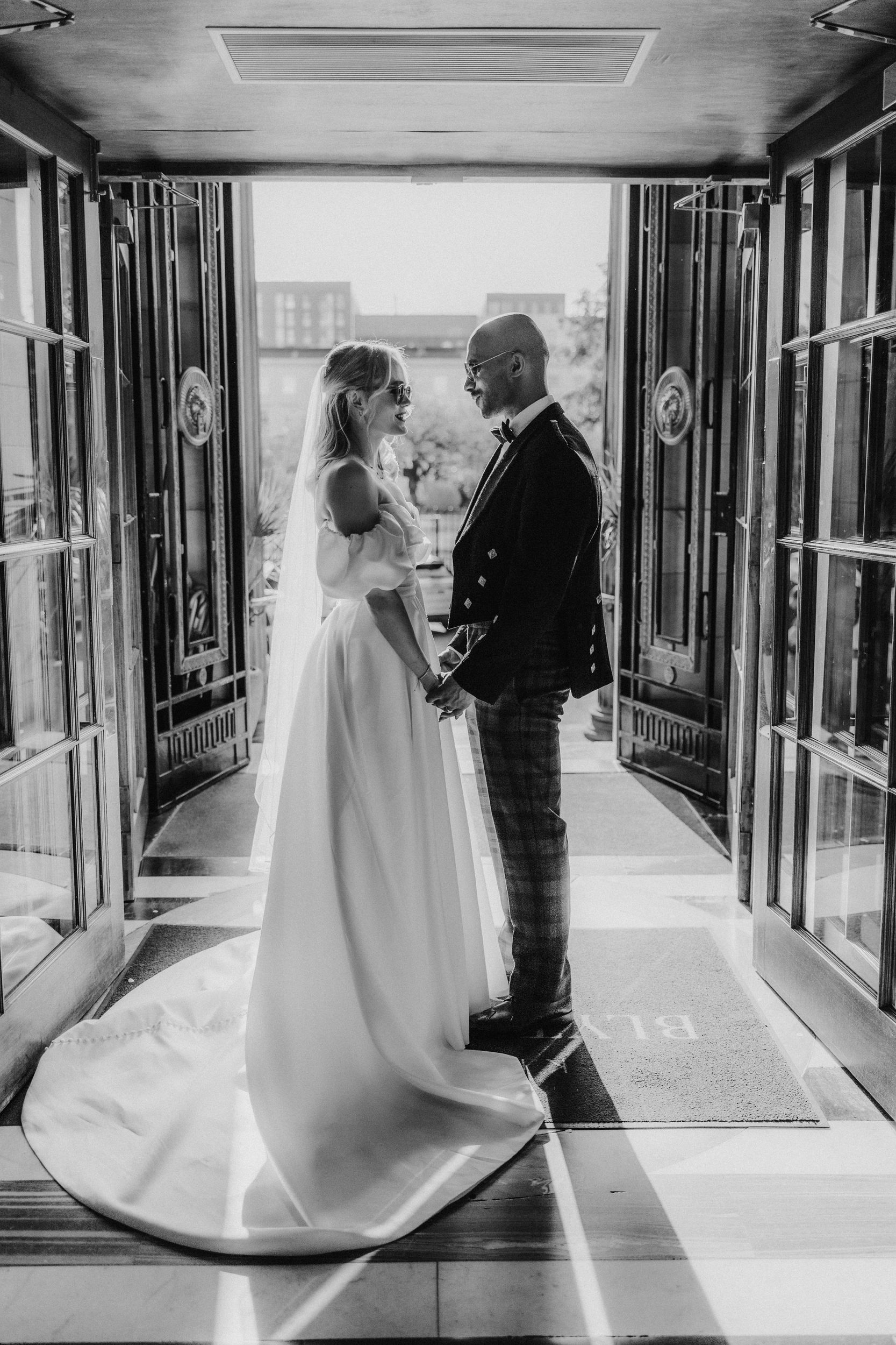 Bride and groom holding hands and facing each other in the open entrance of Kimpton Blythswood Square, backlit, black-and-white.