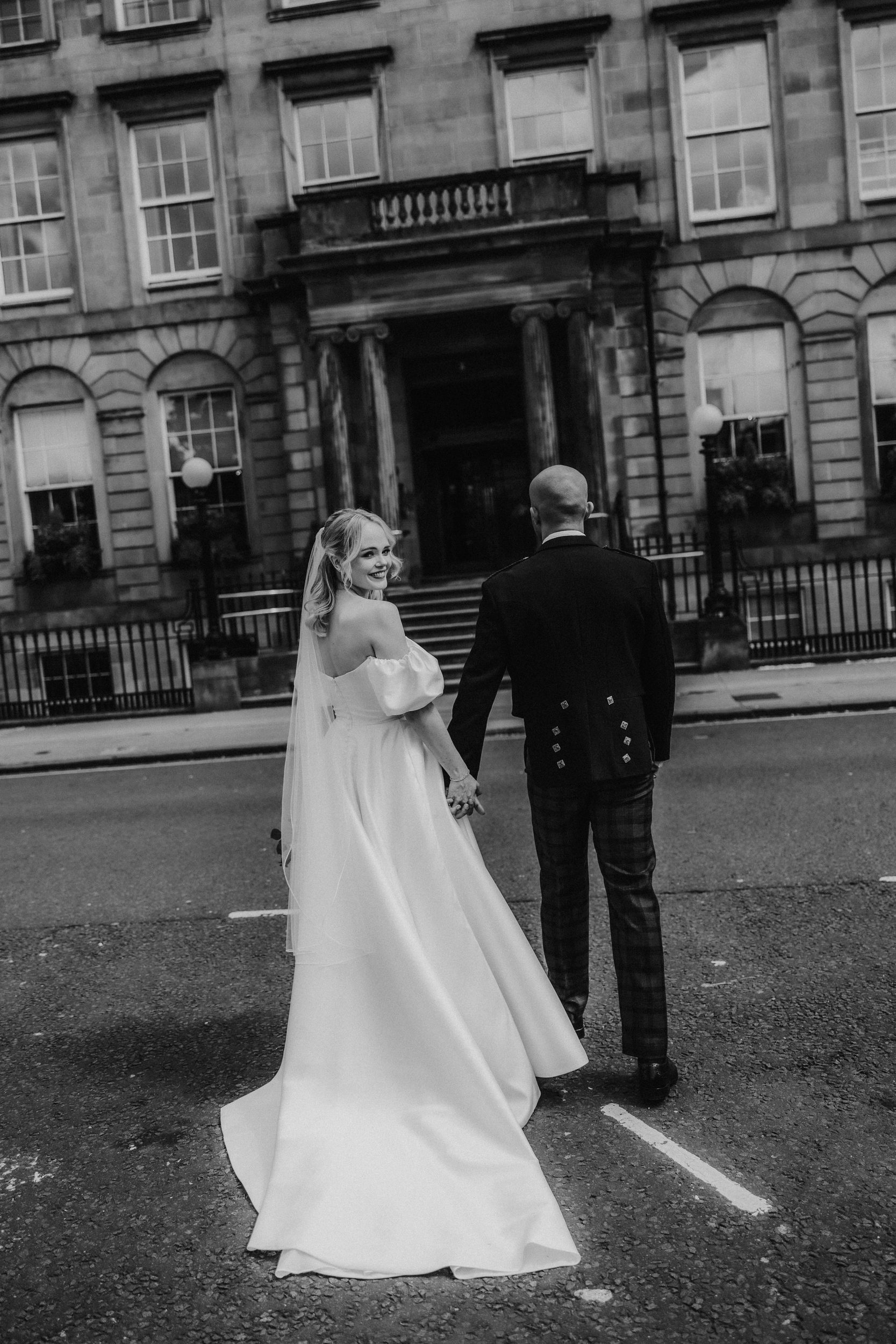 Bride and groom holding hands outside a grand stone hotel as the bride looks back and smiles.