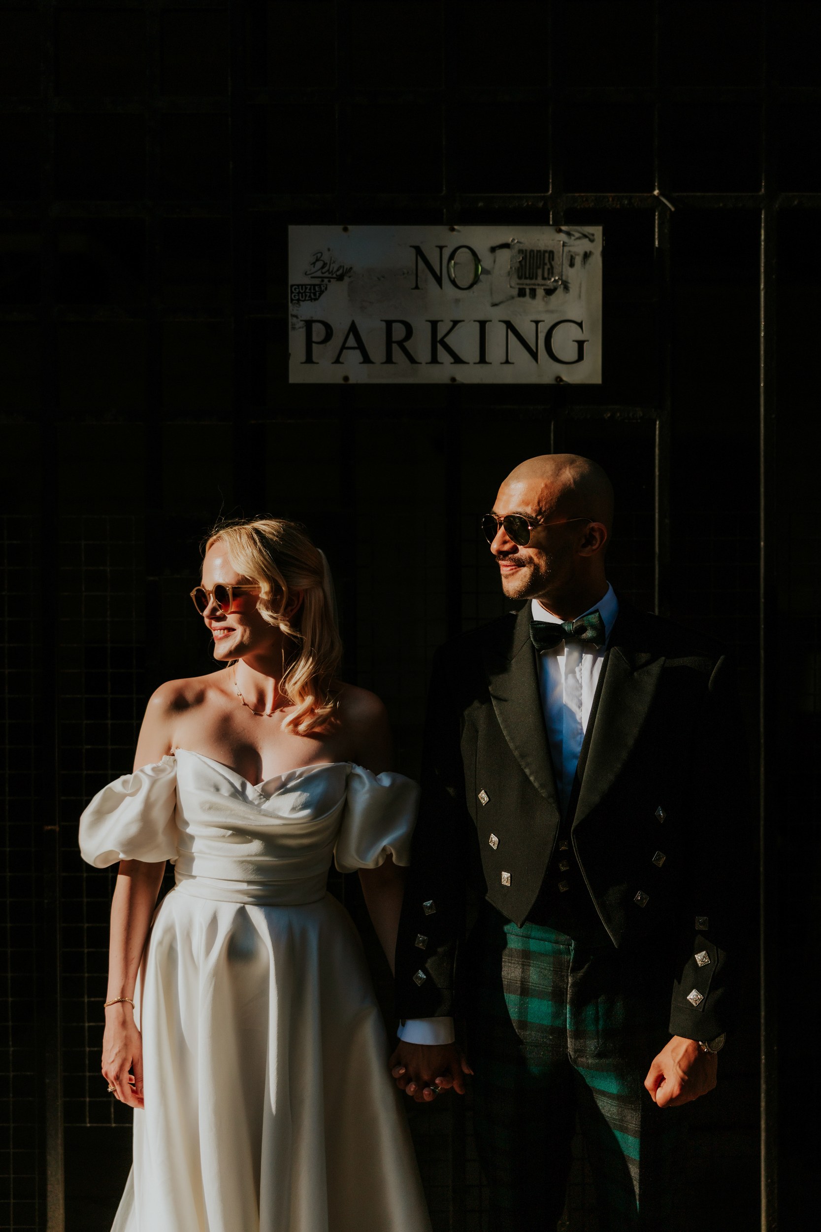 Bride and groom in wedding attire and sunglasses holding hands beneath a 'No Parking' sign.