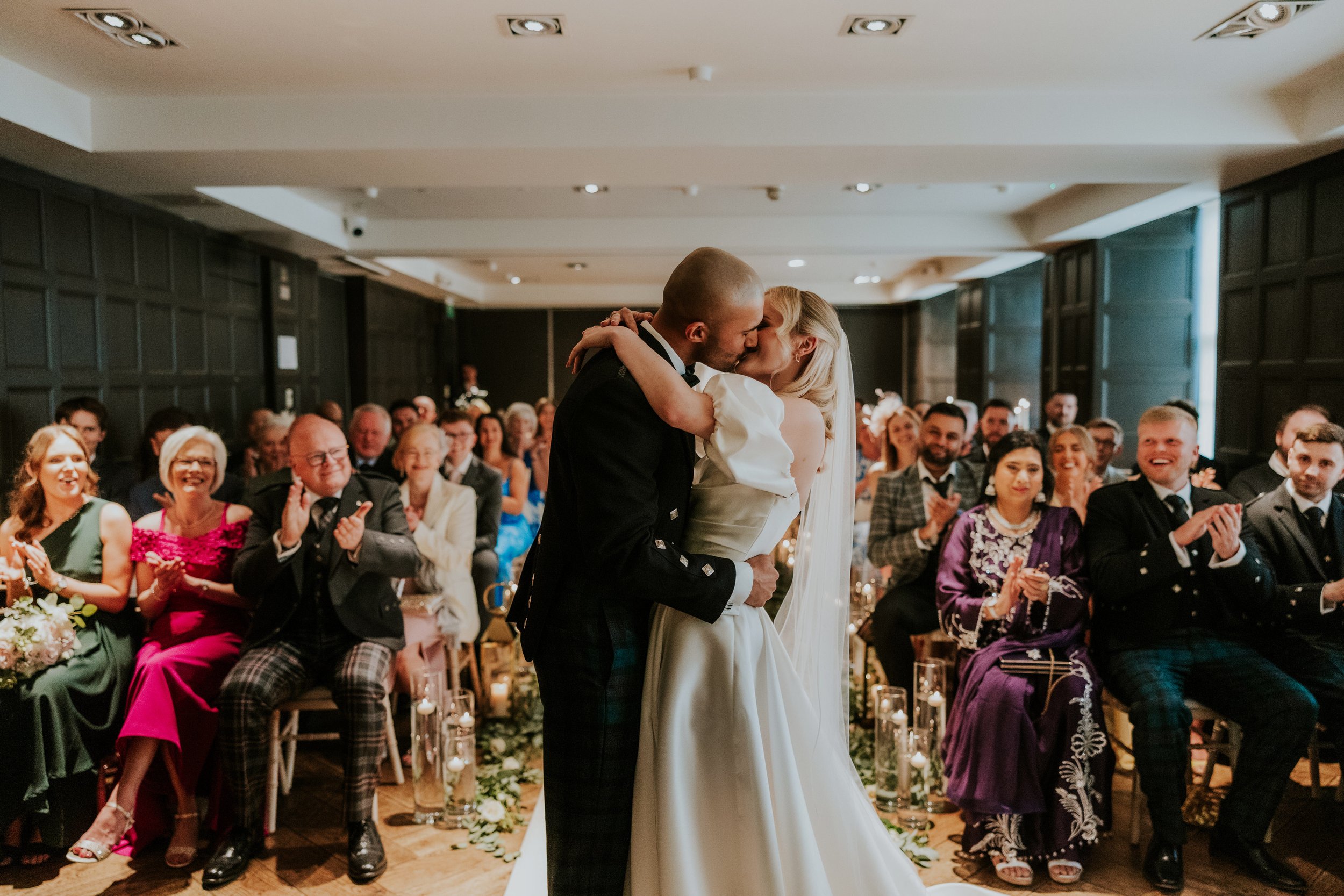 Bride and groom kiss at the end of their ceremony as guests applaud in a candlelit aisle at Kimpton Blythswood Square.