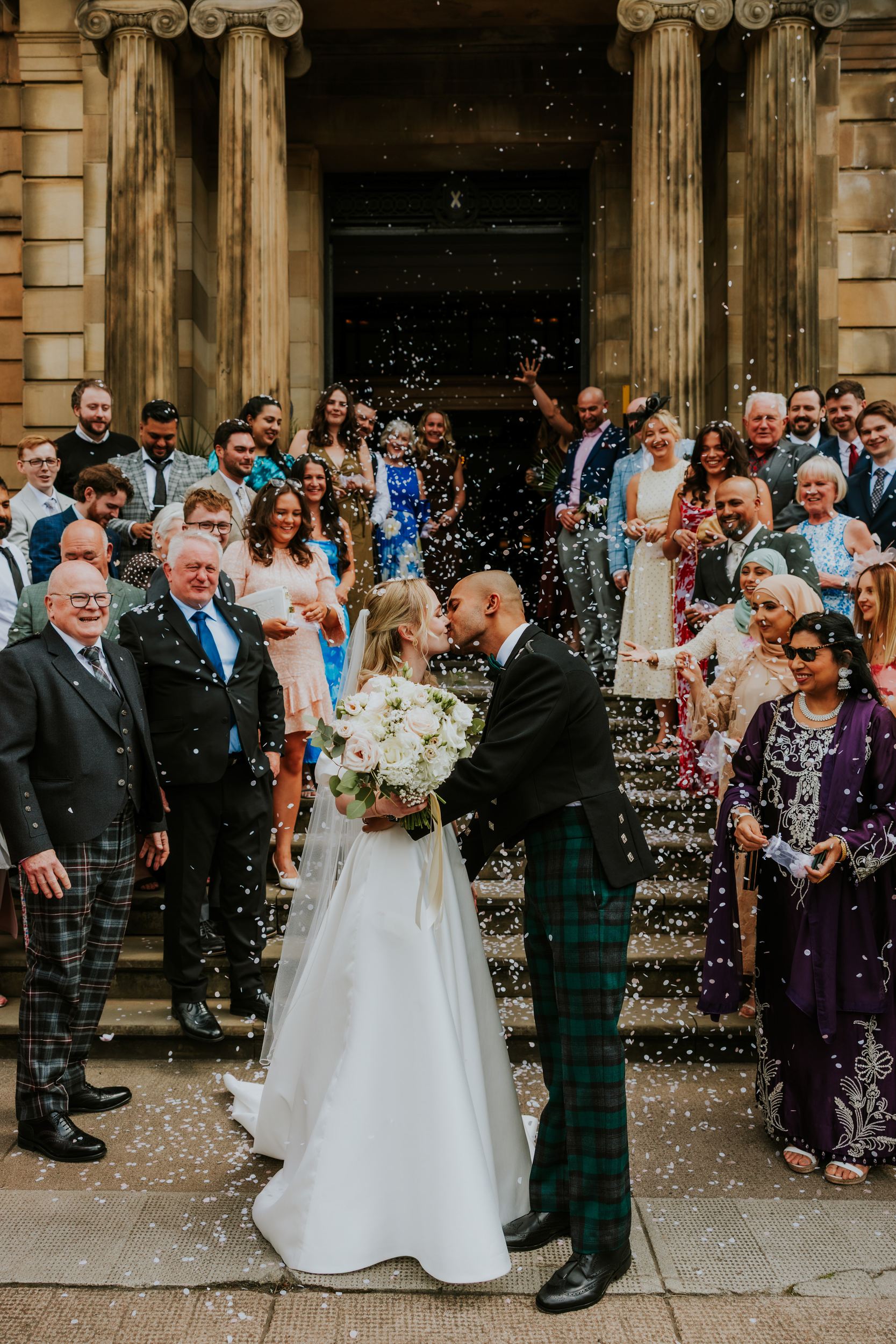 Bride and groom kiss on steps as guests throw confetti; bride with bouquet, groom in tartan trousers.