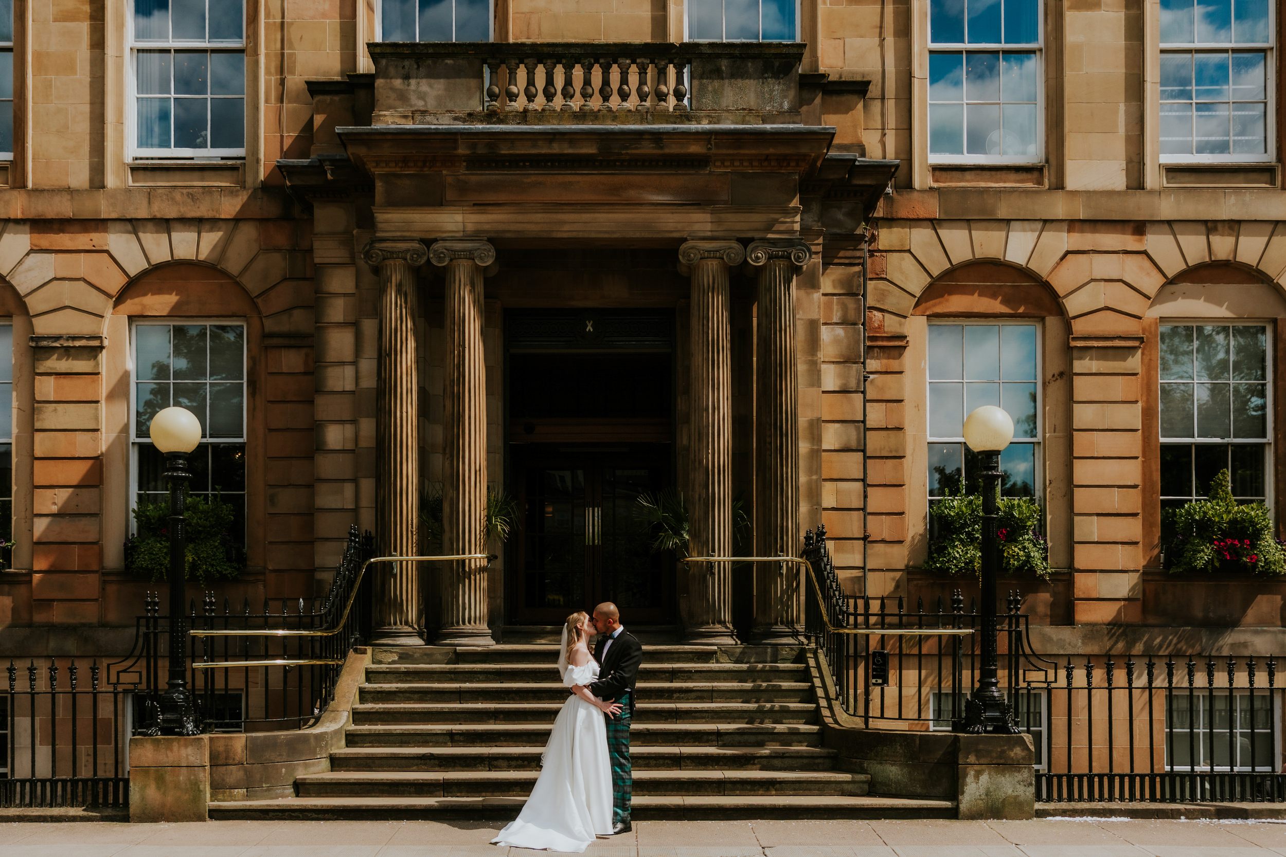 Bride and groom kissing on stone steps outside a grand hotel entrance with columns and sash windows.