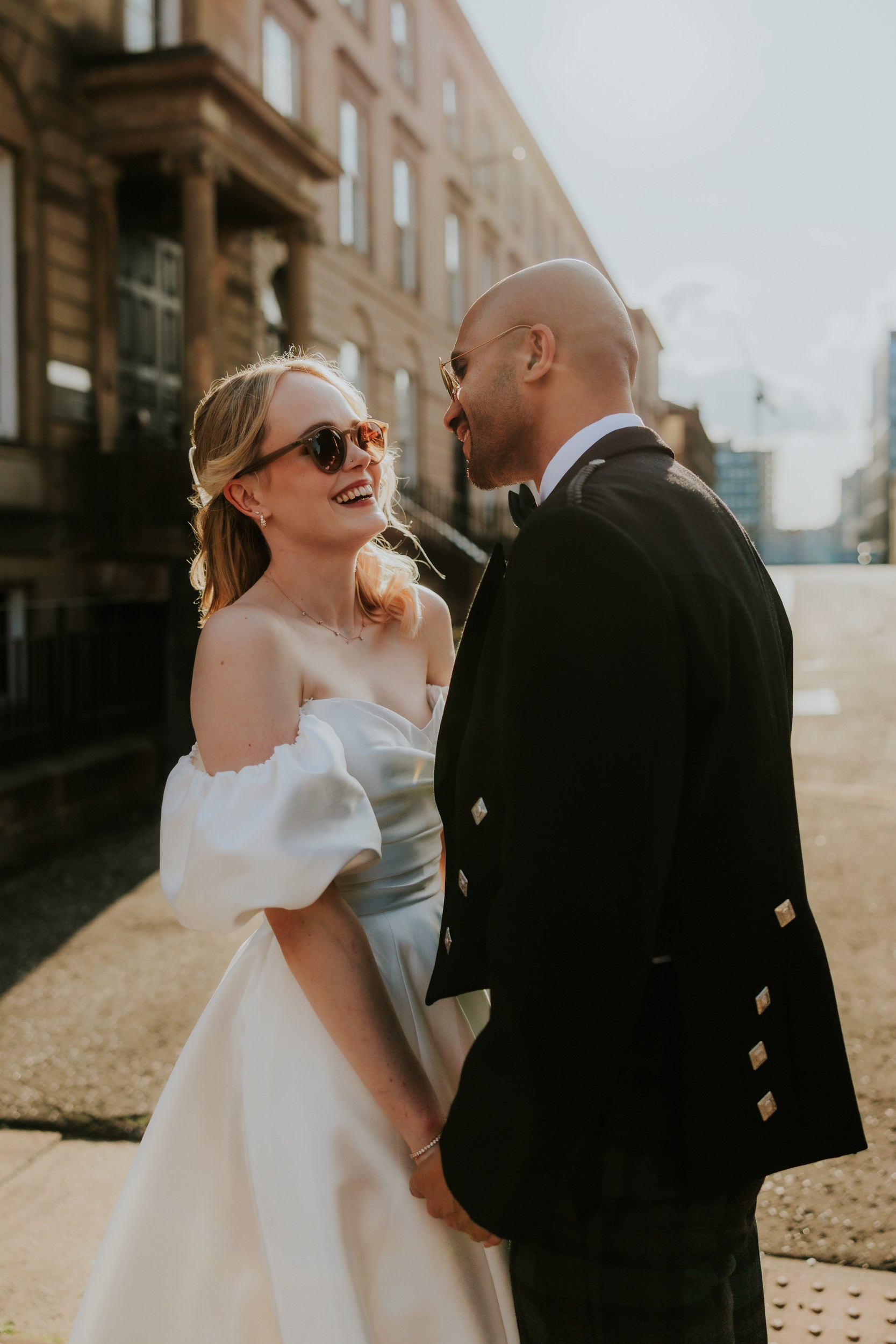 Bride and groom laughing and holding hands on a sunlit street outside a stone building.