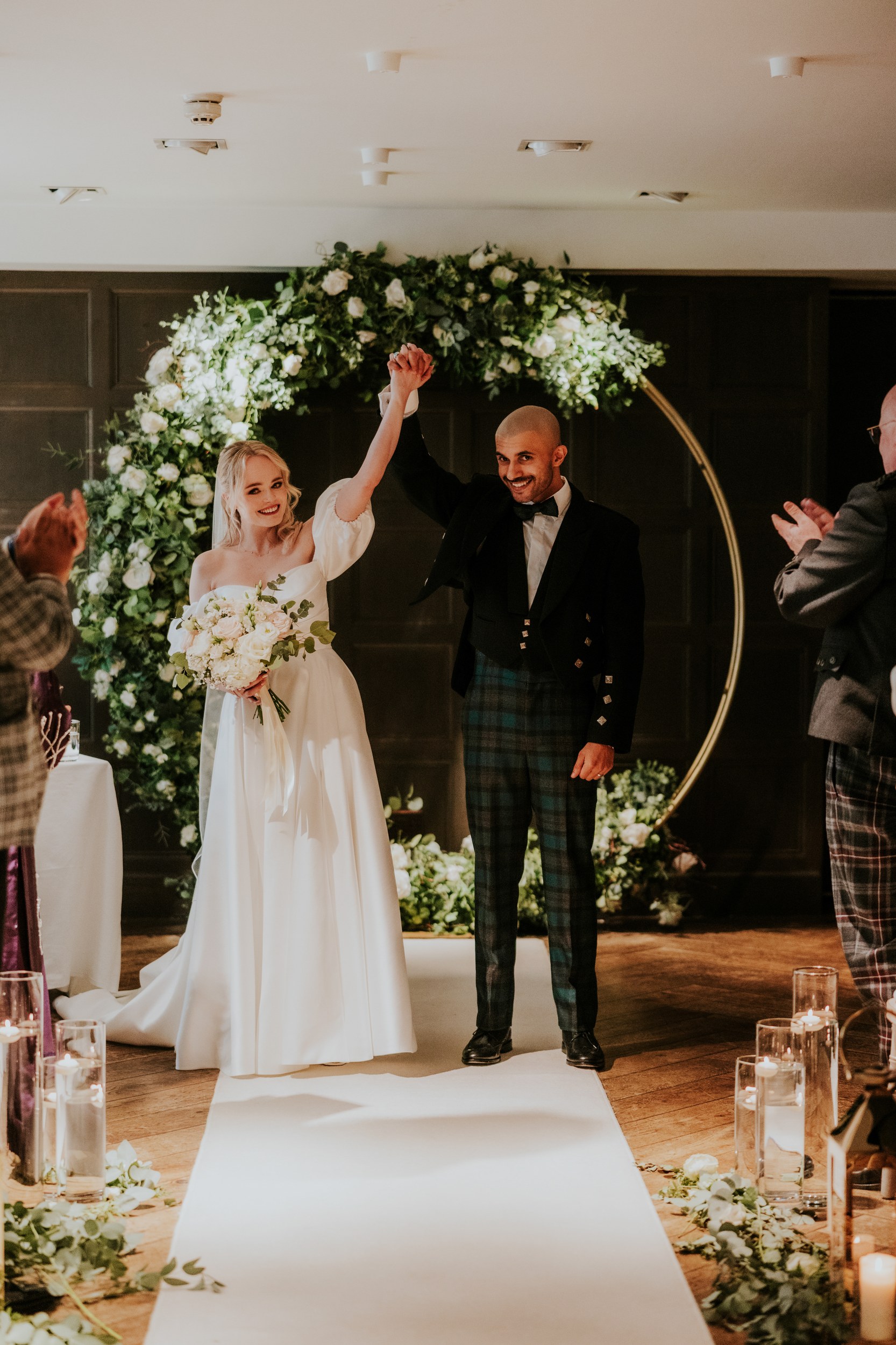 Bride and groom raise joined hands under a floral arch at their indoor ceremony, bride holding a bouquet while guests applaud.