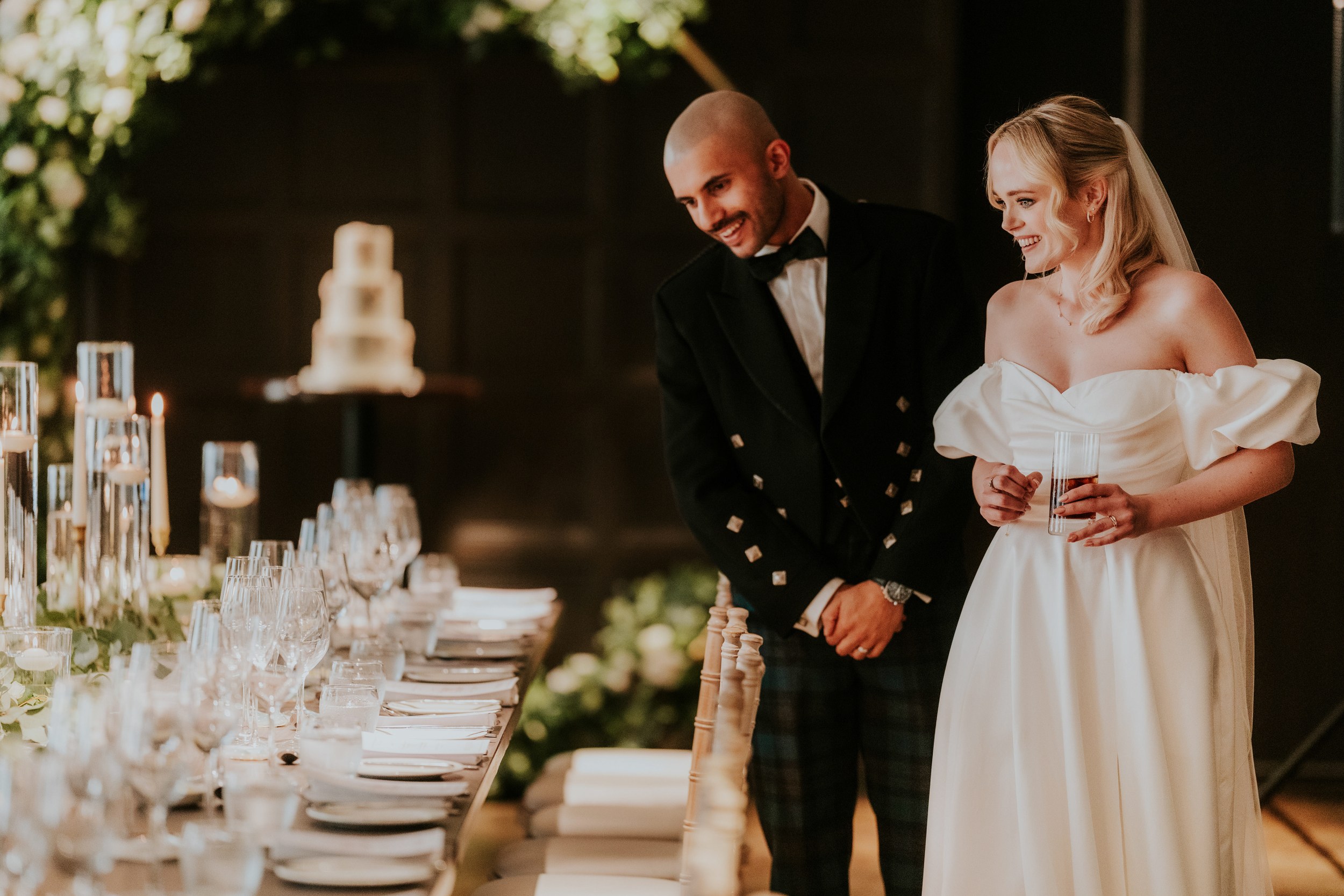 Bride and groom smiling beside a candlelit reception table with a blurred tiered wedding cake in the background.