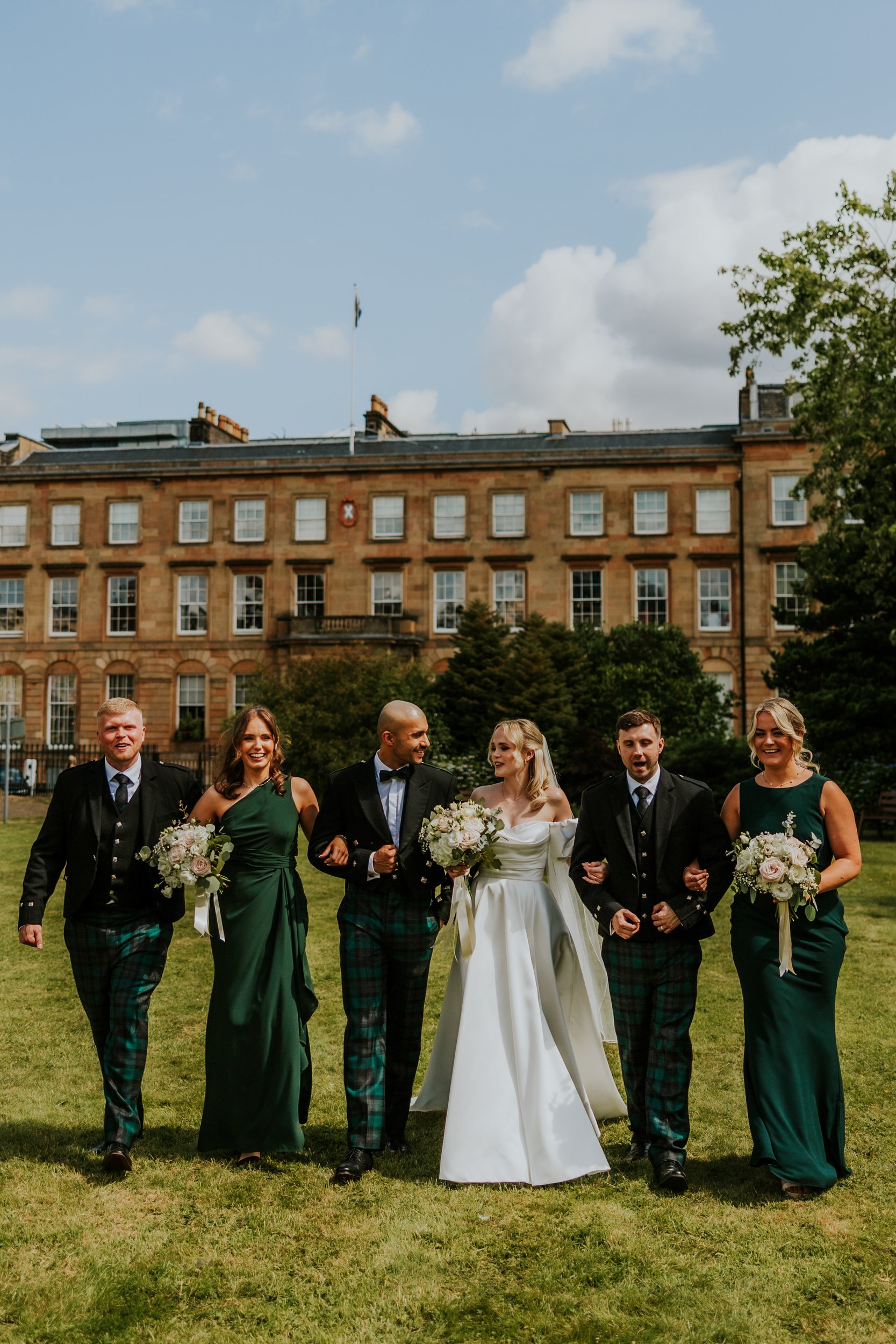 Bride and groom with bridesmaids in green and groomsmen in tartan kilts walking on a lawn in front of a stone hotel.
