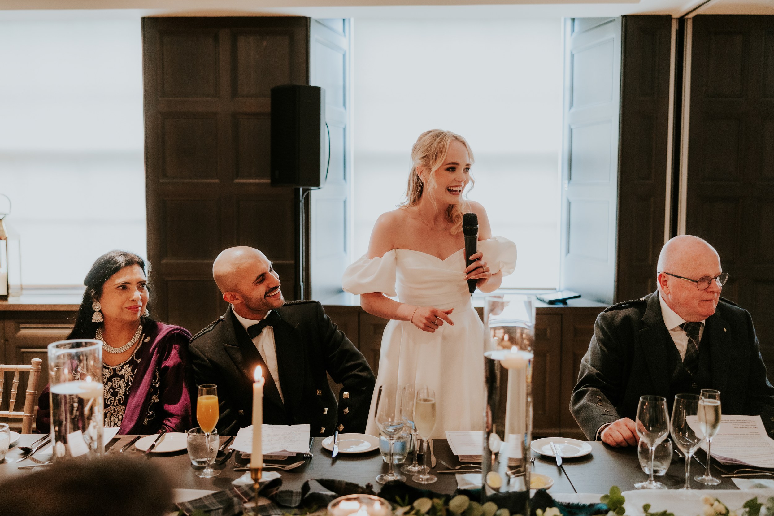 Bride in a white dress speaking into a microphone at the top table while the groom and guests listen.