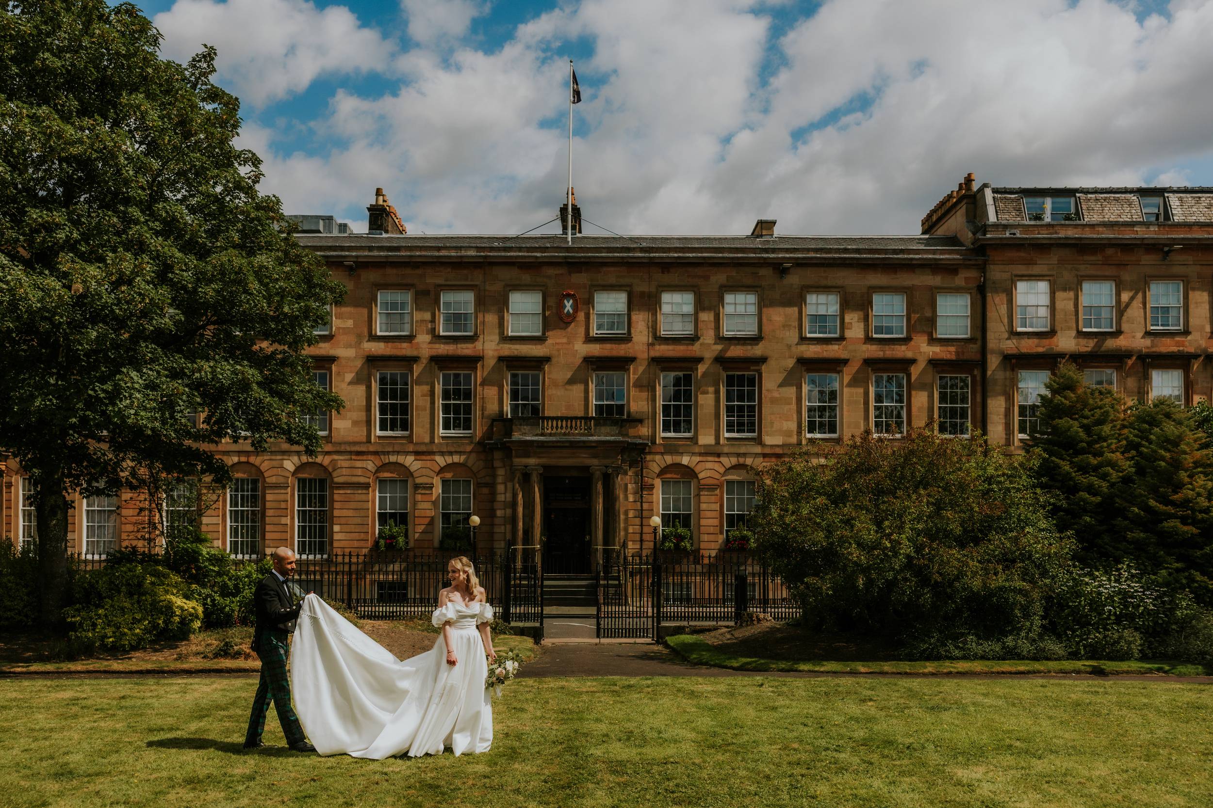 Bride in a white gown and groom holding her train on the lawn outside Kimpton Blythswood Square.