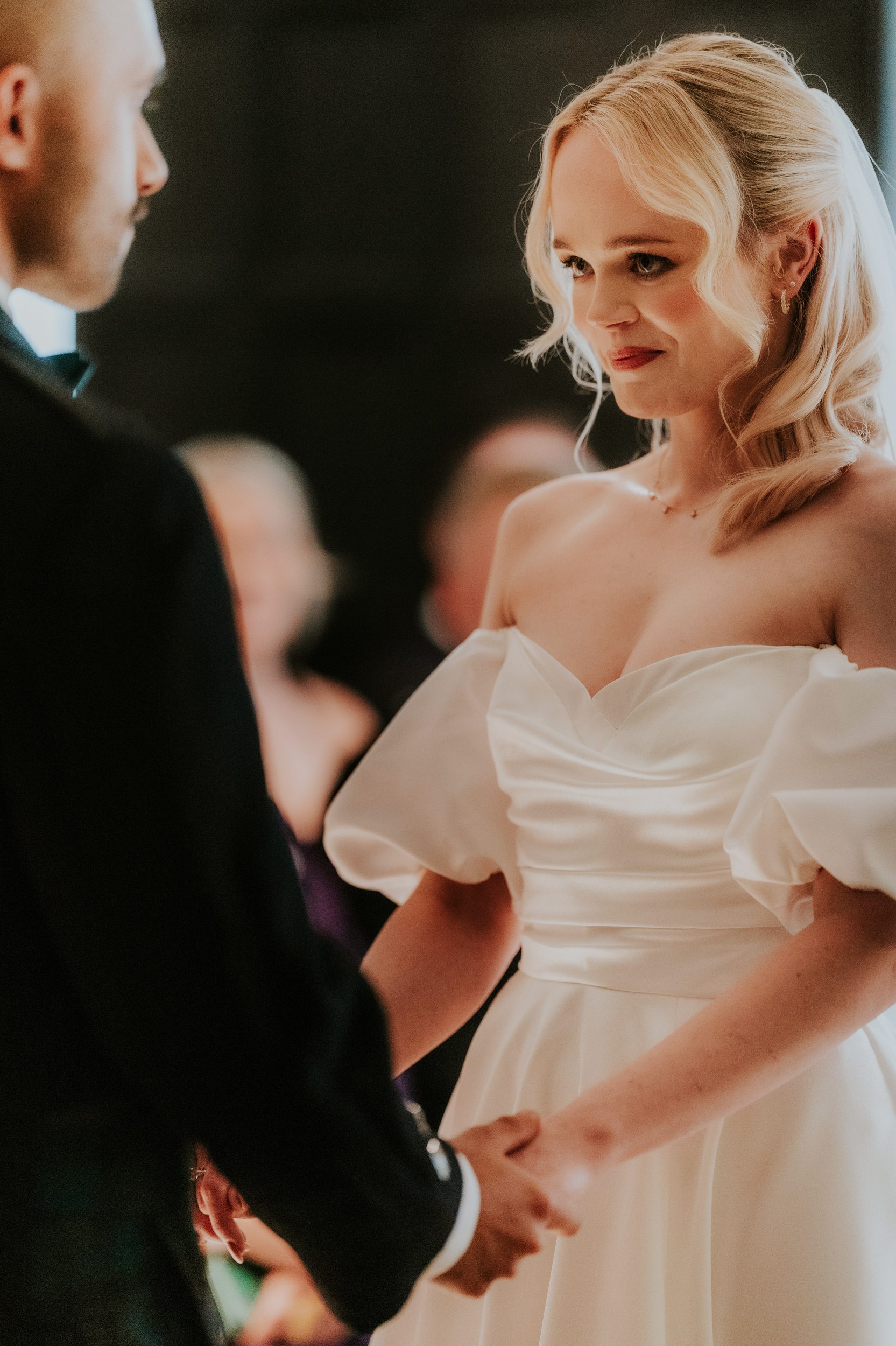 Bride in an off-shoulder wedding dress smiling at her partner as they hold hands during the ceremony.