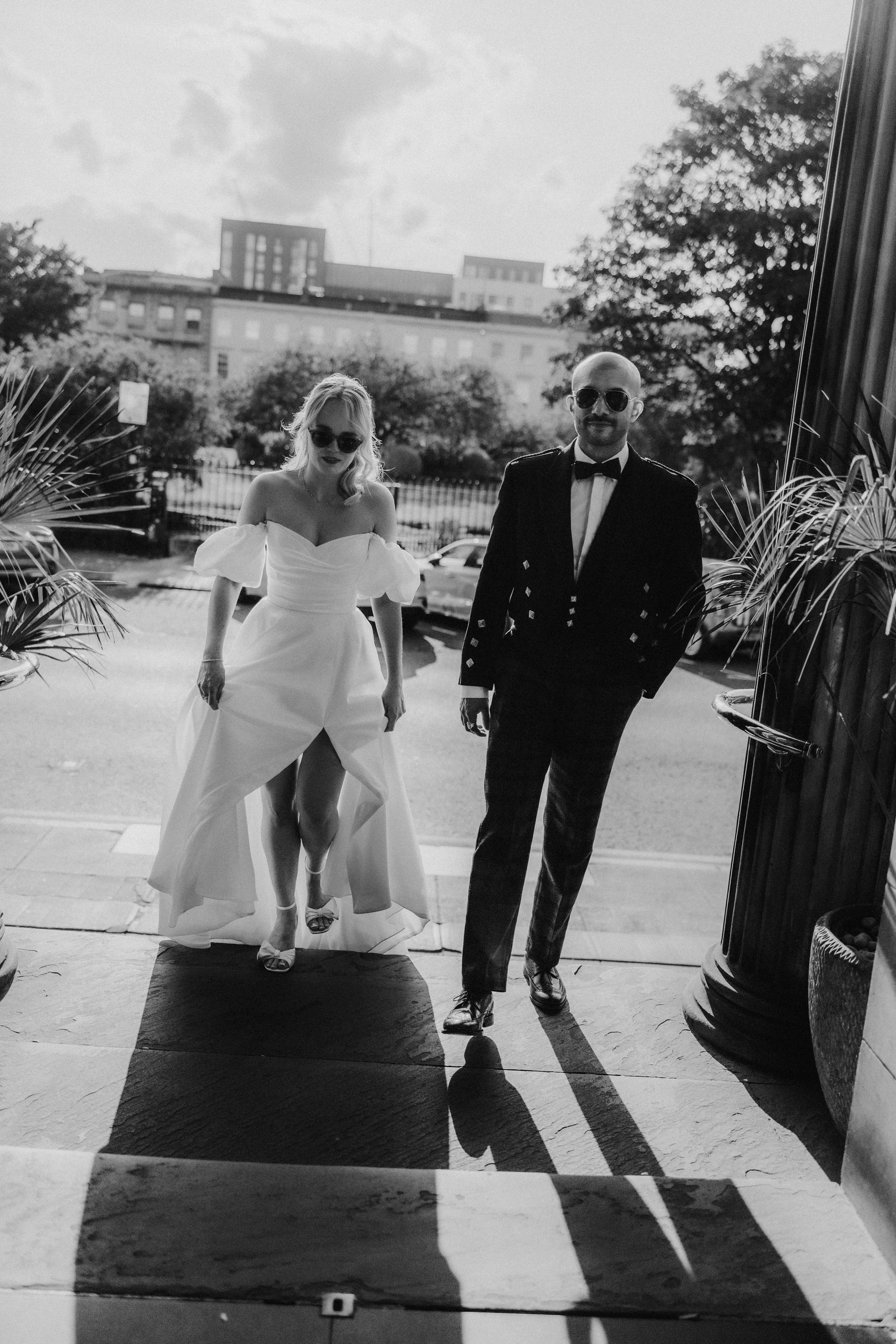Bride in off-shoulder gown and groom in formal jacket, both wearing sunglasses, walking into Kimpton Blythswood Square.
